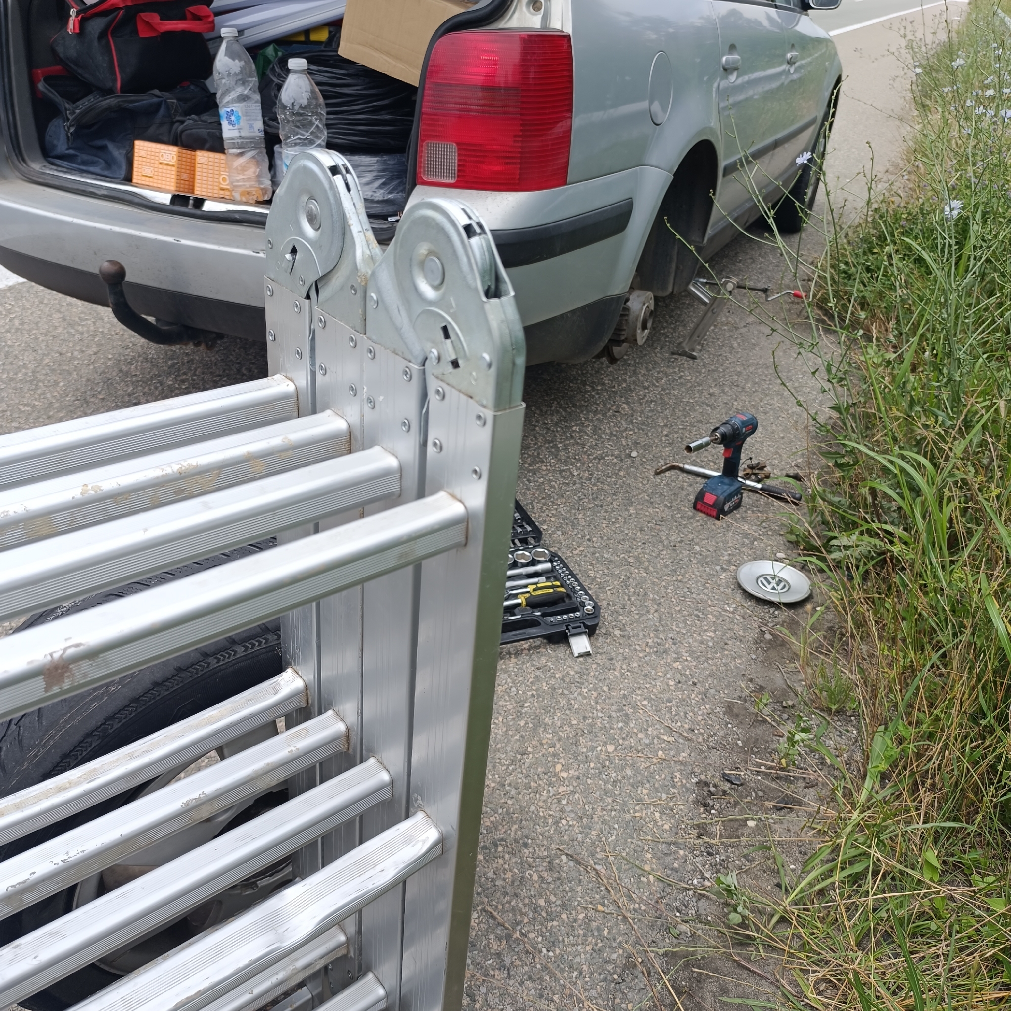 The image shows a silver car parked on the side of a road, likely in a rural area given the presence of tall grass and weeds along the roadside. The car's trunk is open, revealing various items including two plastic water bottles, a black bag, and some orange boxes. The rear passenger-side wheel is missing, and a jack is visible underneath the car, suggesting that a tire change is in progress or has just been completed. A silver ladder, possibly for transport, is leaning against the car and extends into the foreground, obscuring part of the vehicle and the tire area.
On the ground near the car, tools are laid out, including a toolbox containing sockets, a yellow-handled tool, and a cordless drill with a socket attached. A silver Volkswagen hubcap is also on the ground. The scene appears to be taking place during daylight, with neutral weather conditions. The overall impression is one of a car breakdown or preparation for a task requiring the use of the ladder and tools, with the owner likely attending to the car. The location is identified as Krum, Bulgaria.