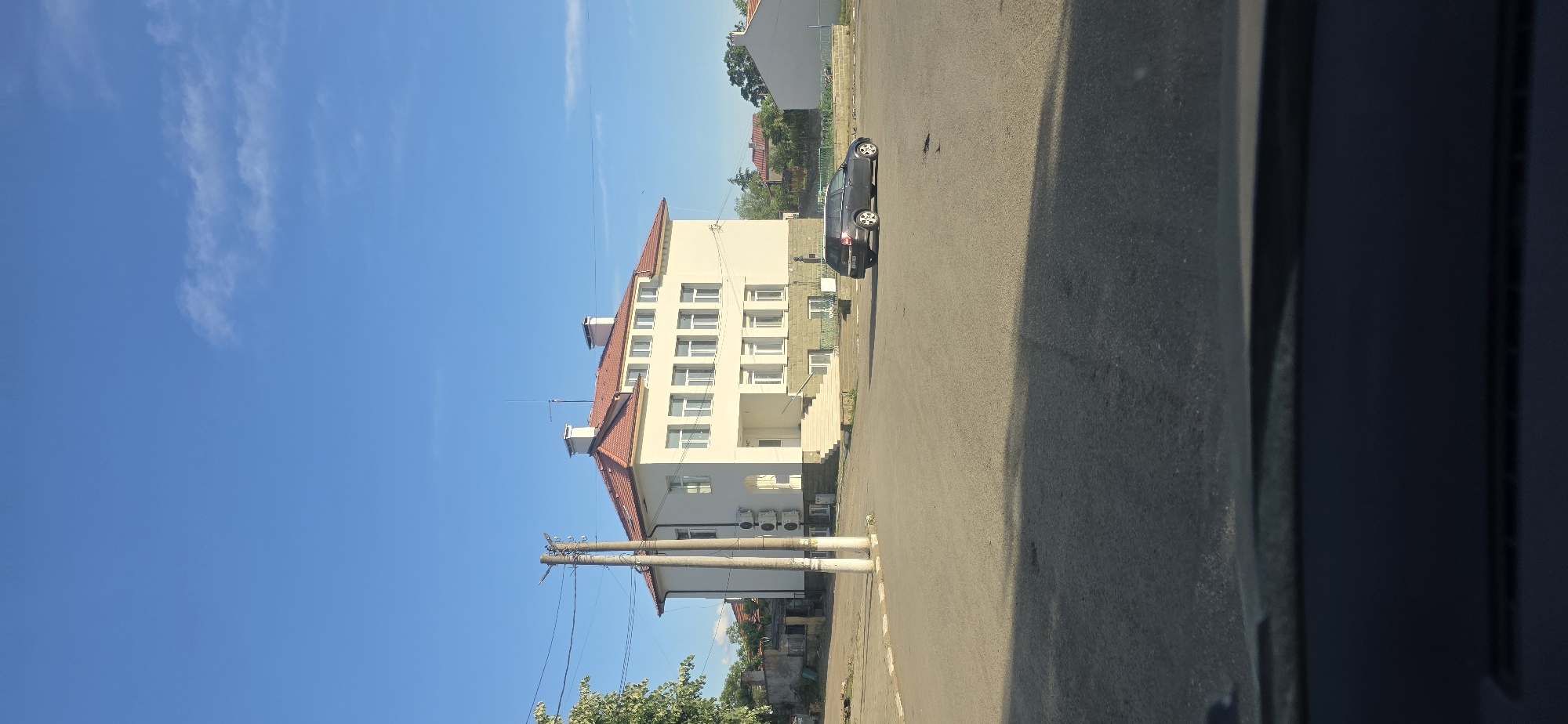 The image captures a street scene in Troyanovo, Bulgaria. A light-colored, multi-story building with a red roof is prominent on the left, situated next to a dark gray car parked on the side of the road. Utility poles and wires run alongside the building. The sky is clear blue with some sparse clouds. A portion of the road is visible, partially shaded by the camera's perspective. A gray panel is visible on the right edge of the image.