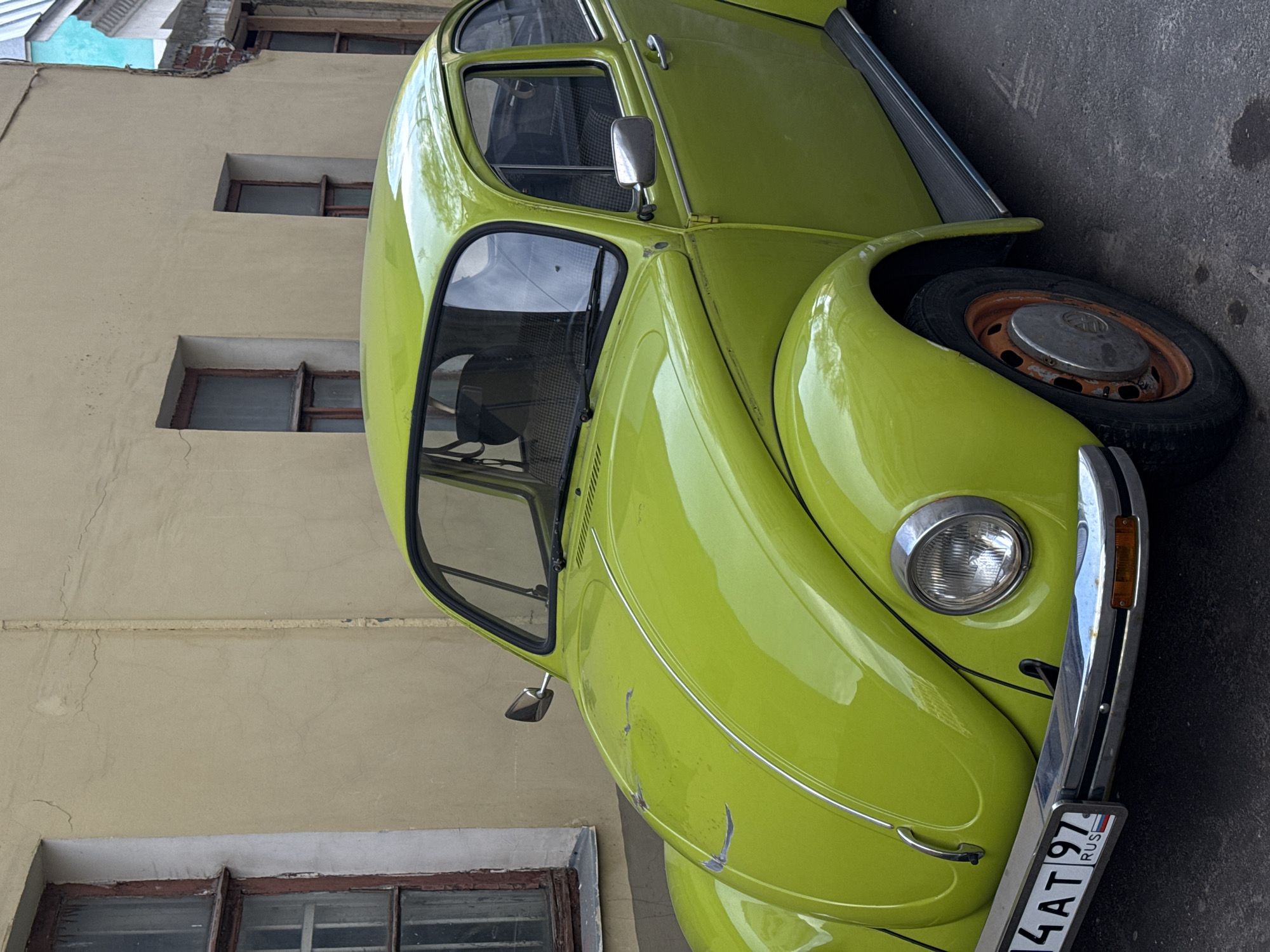 The image shows a lime green Volkswagen Beetle parked on a street in Moscow, Russia. The car is positioned alongside a light yellow building with brown-framed windows. The car's license plate is visible and contains Russian text. The street is asphalt.