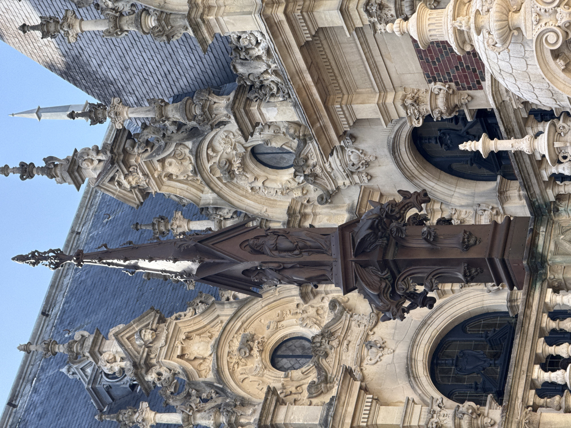 An upward-angled, detailed shot captures the upper facade and roofline of an ornate historical building, likely in Fécamp, France. The dominant feature is a steeply pitched roof covered in dark, diamond-patterned slate tiles, visible predominantly in the upper left and center.
Piercing the clear blue sky, a tall, dark, intricately designed metal spire stands prominently in the center, adorned with what appear to be carved figures or statues within its structure. To its left, a lighter stone pinnacle with decorative elements also extends upwards.
Below the roofline, the building's facade is a rich tapestry of elaborate stonework. It features numerous carvings, including swirling scrolls, gargoyles, and other sculptural details, showcasing a complex architectural style with Gothic and Renaissance influences. Several recessed windows are visible, including circular and arched openings, some fitted with dark grilles or stained glass. Portions of ornate stone balustrades and decorative columns further enhance the facade's intricate design. The light-colored stone shows signs of weathering, indicating the building's age. The scene is illuminated by bright daylight, suggesting a clear day. No human activity or visible text is present.