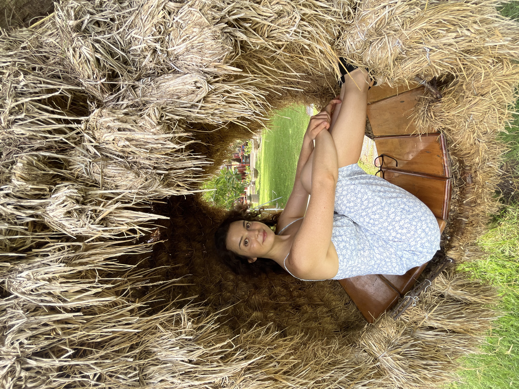 A woman with curly brown hair and fair skin is seated inside a structure made of dried hay. She is wearing a light blue dress with a floral pattern. Her legs are crossed, and she is leaning back, resting her arms on her legs. She is looking towards the camera and smiling. The hay structure forms a hollow space, with the dried stalks hanging down in front of her. Through an opening in the hay, a green lawn and some trees are visible in the background. The overall scene suggests an outdoor setting, possibly a park or a resort in Mũi Né, Vietnam.