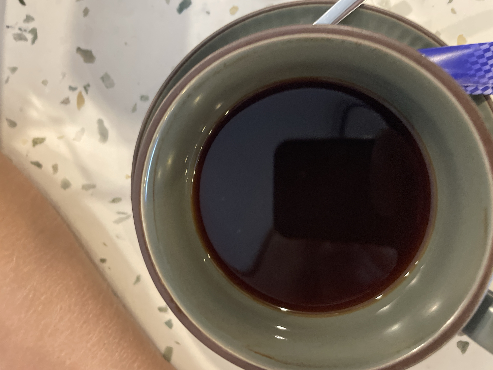An overhead shot shows a cup of black coffee on a terrazzo table in what appears to be a cafe in Bac Nha Trang Ward, Vietnam. The coffee is dark and still, with a slight reflection on its surface. The cup is made of a ceramic material, with a green exterior and a brown rim. A silver spoon rests on the saucer, next to a small blue and white packet, possibly containing sugar or creamer. A person's arm is visible in the lower left corner of the frame, suggesting they are holding or near the cup. The terrazzo table has a white background with scattered grey, green, and yellow flecks.