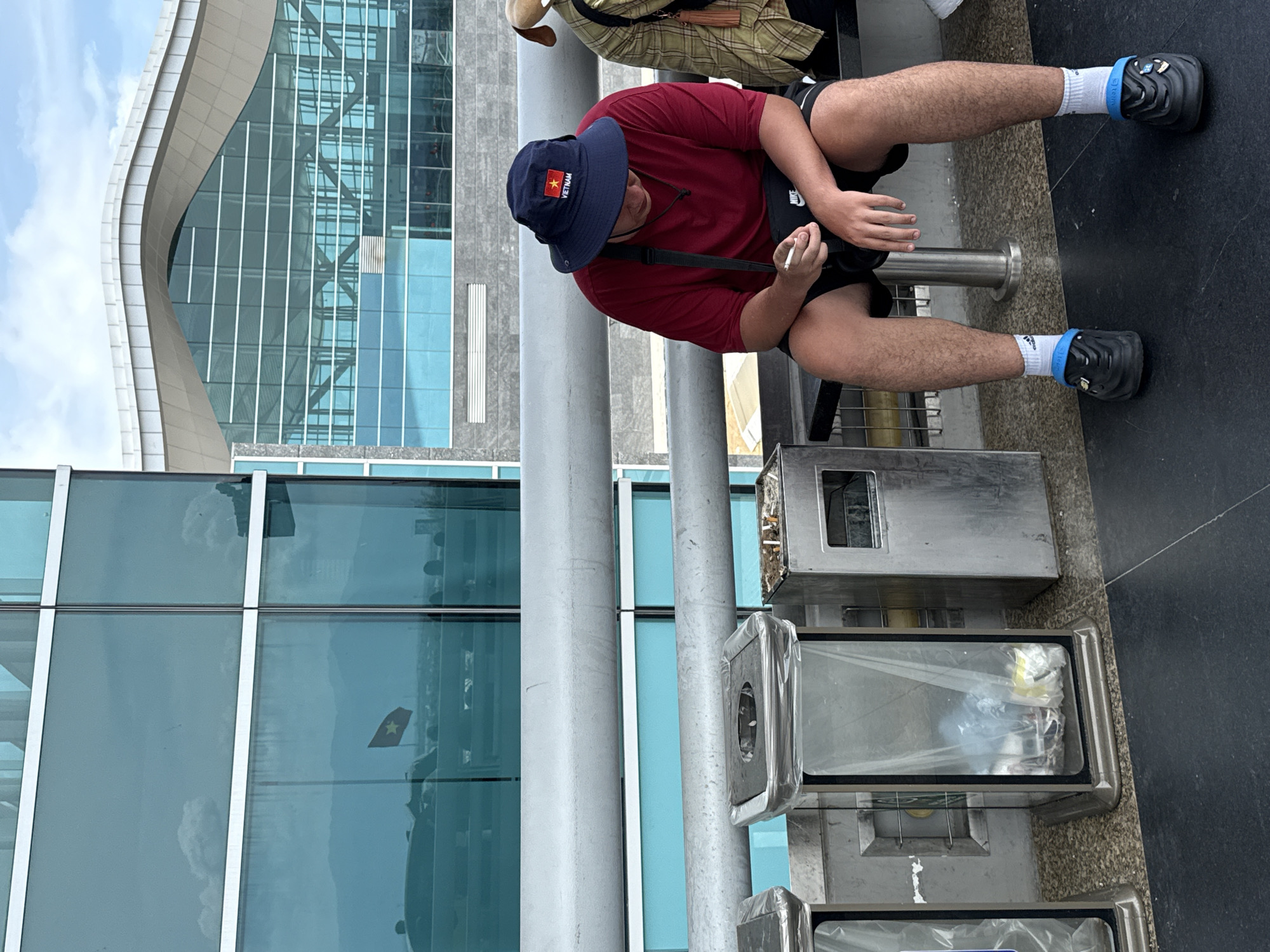 A man wearing a red shirt, black shorts, and a navy blue bucket hat with the Vietnamese flag is sitting outdoors, holding a cigarette. He is positioned near a series of metal trash receptacles. The background features a modern building with a glass facade reflecting the sky and a Vietnamese flag visible in the reflection. The scene appears to be in Bac Cam Ranh Ward, Vietnam.