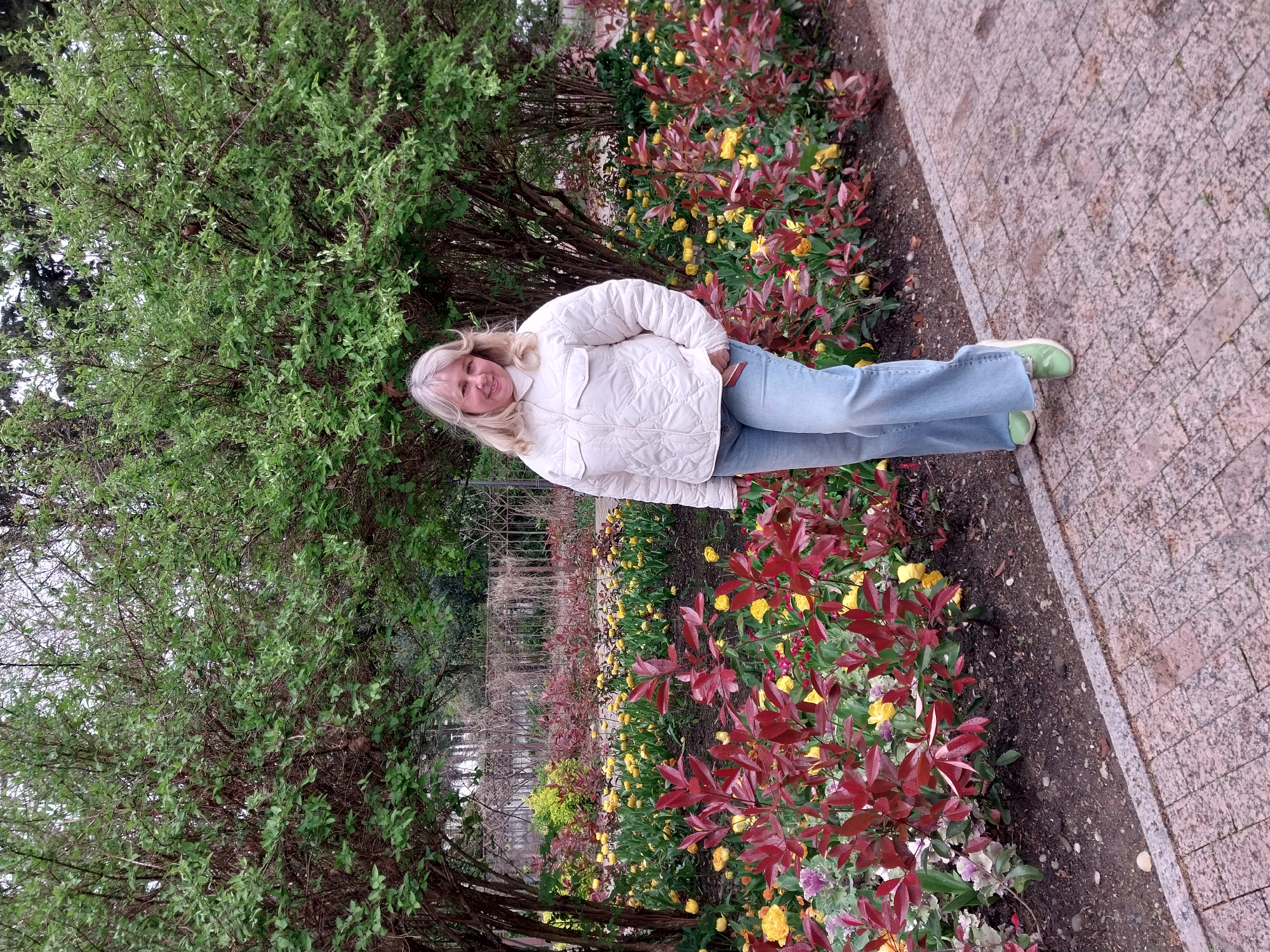 A medium shot shows a woman standing alongside a pathway in a garden. The garden is blooming with yellow and red flowers, and green shrubbery is abundant. The pathway is made of reddish-brown bricks. The woman is fair-skinned, with blonde hair and is wearing a white jacket, blue jeans, and light green shoes. The garden may be located in the city of Sochi, Russia.