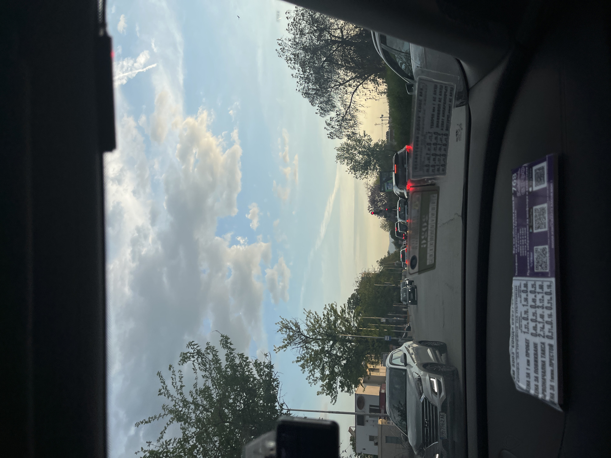 The image, taken from inside a car, shows a street scene in Plovdiv, Bulgaria. The car is in traffic, with other vehicles visible ahead. A car, with license plate PB7962AE, is visible on the right side of the car taking the picture. Trees line the road, and the sky is partly cloudy. There are stickers/tickets affixed to the car's dashboard. The OCR reveals some price listings for taxi services.