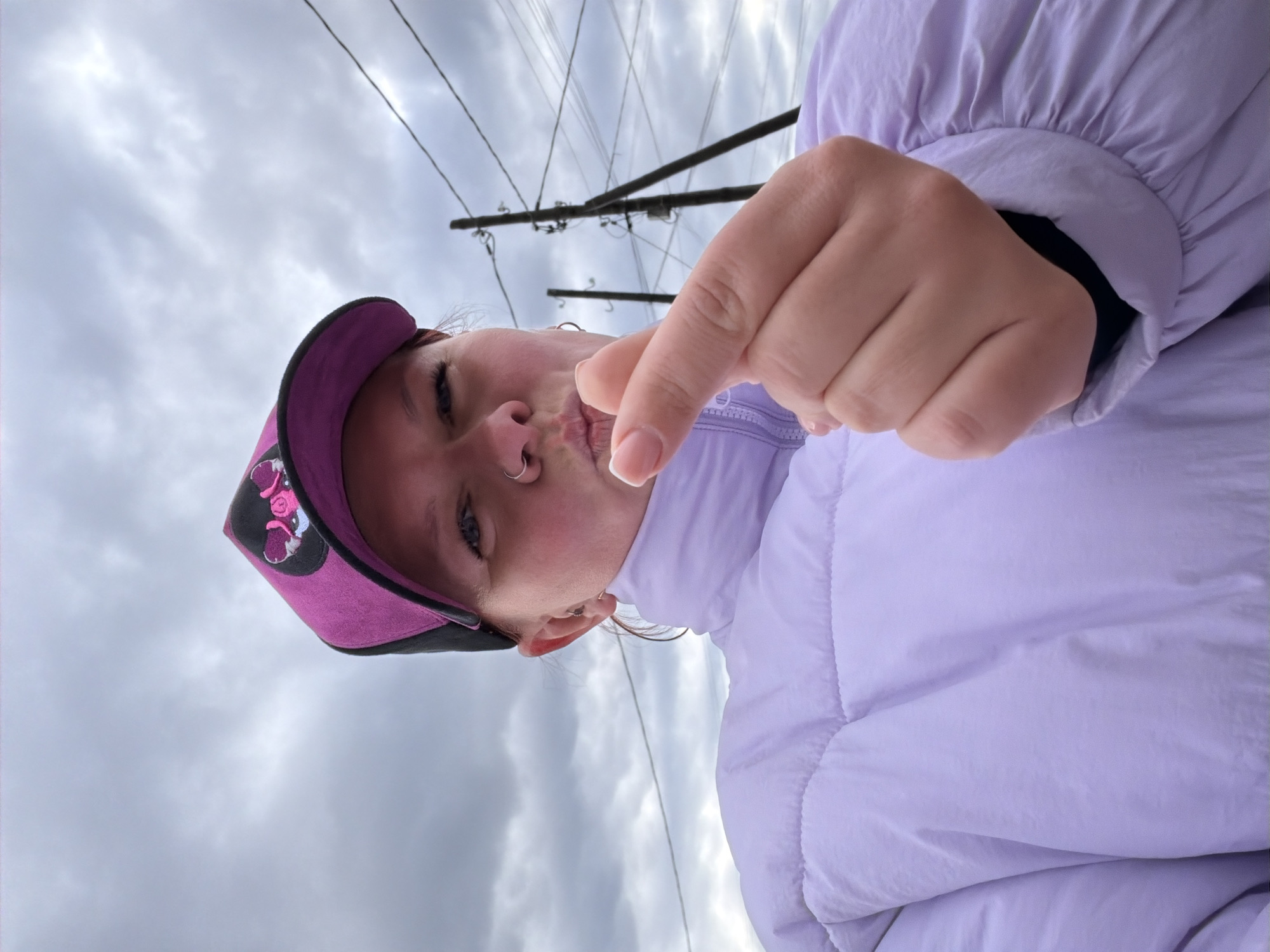 The image captures a low-angle selfie of a person outdoors in Chaikovsky, Russia, on an overcast day. The subject, likely female, is wearing a light purple padded jacket and a purple and black baseball cap featuring an embroidered design that appears to be a stylized head or logo with pink elements. She has a nose piercing (a hoop) and an earring visible. Her face is angled slightly upwards, with one eye open and her lips pursed in a playful expression. Her right hand is extended towards the camera, forming a finger heart gesture with her thumb and forefinger.
The background shows a cloudy, grey sky, indicating an overcast or possibly cold day, which aligns with the puffy jacket worn by the subject. Utility poles and a network of overhead power lines are visible against the sky, suggesting an urban or suburban outdoor setting. The light is diffused due to the clouds, typical of daytime. No visible text is discernible on the hat or clothing. The overall impression is one of a lighthearted moment captured in a casual outdoor setting.