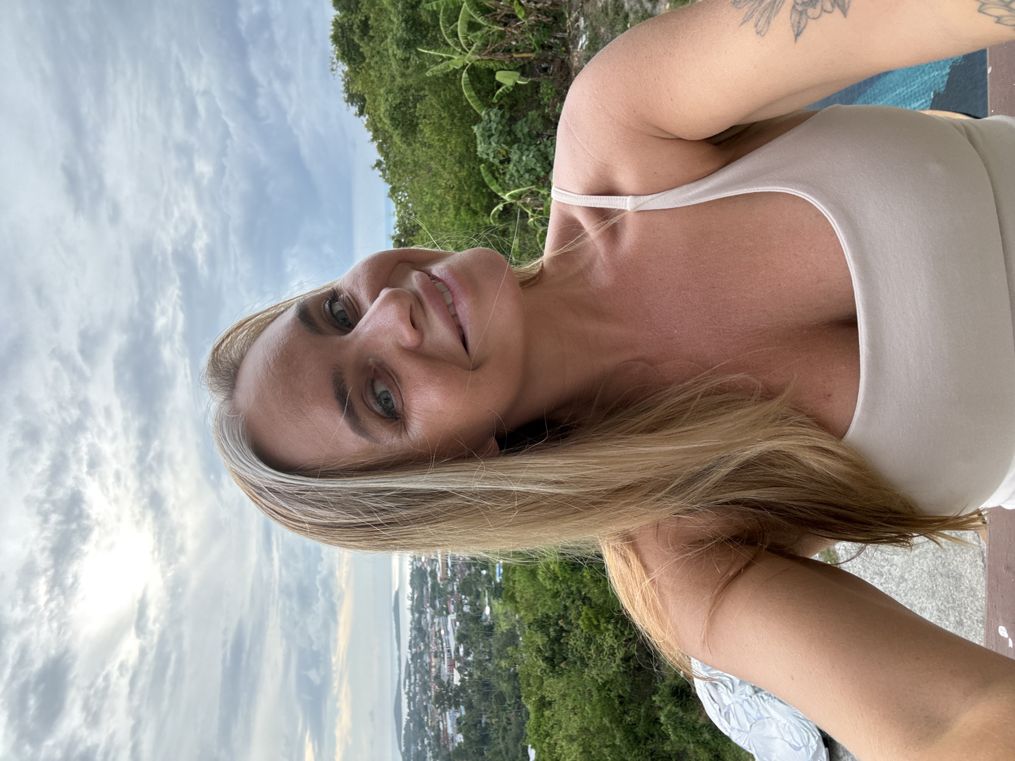 A woman with blonde hair and gray streaks poses for a selfie outdoors. She is wearing a light-colored tank top. Behind her, a city is visible in the distance with the ocean beyond. Lush green trees and banana plants surround the foreground. The sky is overcast with some sunlight breaking through the clouds. The location is Ko Samui District, Thailand.