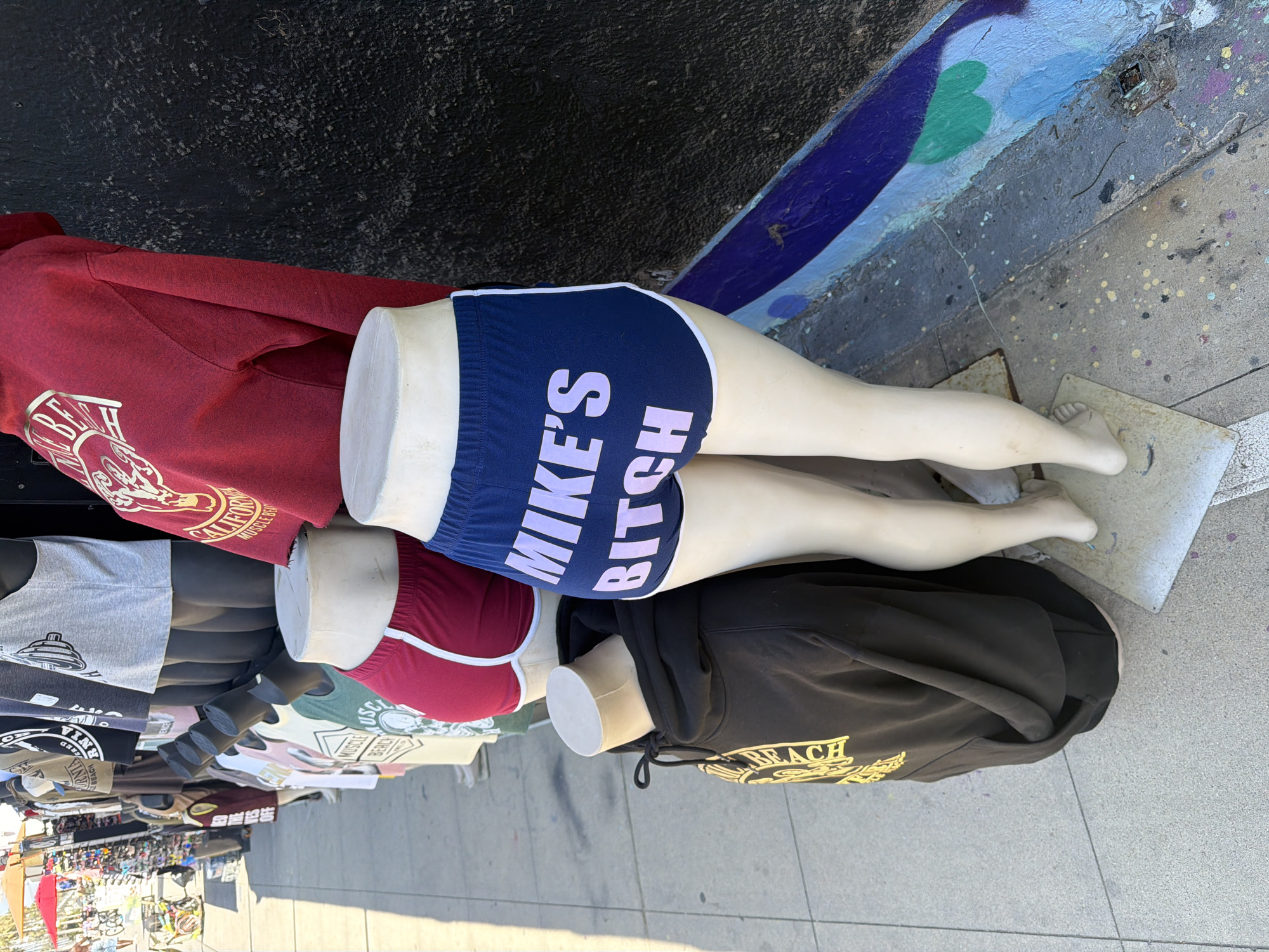 This image captures a portion of an outdoor clothing display, likely at a street stall or market in Los Angeles, USA. The primary subjects are several headless mannequins and torsos adorned with various garments. In the foreground, two pairs of mannequin legs are prominently displayed.
One pair wears navy blue athletic shorts with white trim, which notably feature the bold white text "MIKE'S BITCH" printed on the backside. Below these, another pair of legs wears maroon shorts with white trim. The mannequins' lower legs and feet rest on a concrete sidewalk, which is splattered with colorful paint dots. Behind them, a painted wall or curb features blue and green graffiti.
Further into the display, several mannequin torsos are visible. One wears a red hoodie with text partially visible as "CALIFORNIA MUSCLE BEACH". Another wears a black hoodie with gold text, where "BEACH" is discernible. Other t-shirts and hoodies in the background feature text like "MUSCLE BEACH," "ALIFORN," and other partial words.
The scene is brightly lit by natural sunlight, indicating daytime. Strong shadows are cast on the sidewalk, suggesting a clear, sunny day. The overall setting is typical of an outdoor retail environment, with various merchandise arranged for sale.