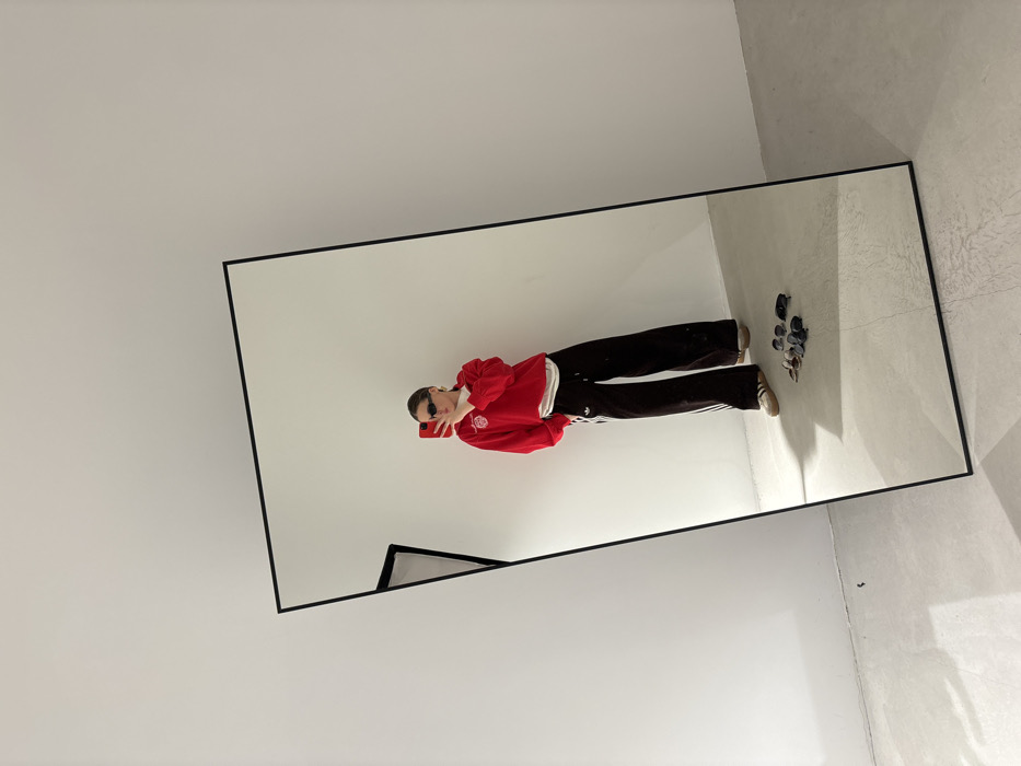 The image displays an indoor scene featuring a person taking a mirror selfie. A large, rectangular mirror with a thin black frame leans against a plain white wall at an angle. The floor is a light grey concrete or similar material. Reflected in the mirror is a female individual wearing a red long-sleeved top with a white collar, a white undershirt, wide-leg black Adidas track pants with three white stripes, and light brown sneakers. She is wearing dark sunglasses and holds a red smartphone in her left hand, aimed at the mirror to capture the reflection. On the floor in front of the mirror, to the right of the person's reflected feet, is a cluster of approximately six to eight pairs of sunglasses. The minimalist environment of white walls and a light floor offers no specific architectural or contextual details to identify a precise location within the city of Krasnodar, Russia.