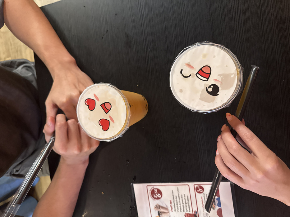 The image depicts a high-angle view of a dark, matte-finish table within a bubble tea shop located in North Miami Beach, United States of America. Two clear plastic cups containing beverages are centrally positioned. The upper cup holds an orange-yellow liquid with dark spherical inclusions, sealed with a plastic film displaying a cartoon face featuring two red heart-shaped eyes, pink blush marks, and an open mouth. The lower cup holds a lighter, opaque liquid, also with dark spherical inclusions, sealed with a plastic film displaying a winking cartoon face with one eye closed, an open mouth showing teeth, and pink blush marks.
In the upper portion of the frame, the forearms and hands of one individual are visible, wearing a dark grey shirt over a light blue layer. Their hands are loosely clasped, resting on the table, and they are holding a metallic reusable straw. In the lower portion of the frame, the left hand of a second individual is visible. This hand has fingernails painted with orange/red tips and holds a black plastic straw, partially encased in its plastic wrapping, positioned near the lower beverage.
A laminated menu is partially visible on the left side of the table. It features red headings and lists "Fruit tea" options such as "Strawberry black tea," "Peach black tea," and "Passion Fruit tea," with a price of "$6.50." Another section for "Popping boba" is partially visible with a price of "$6.75."