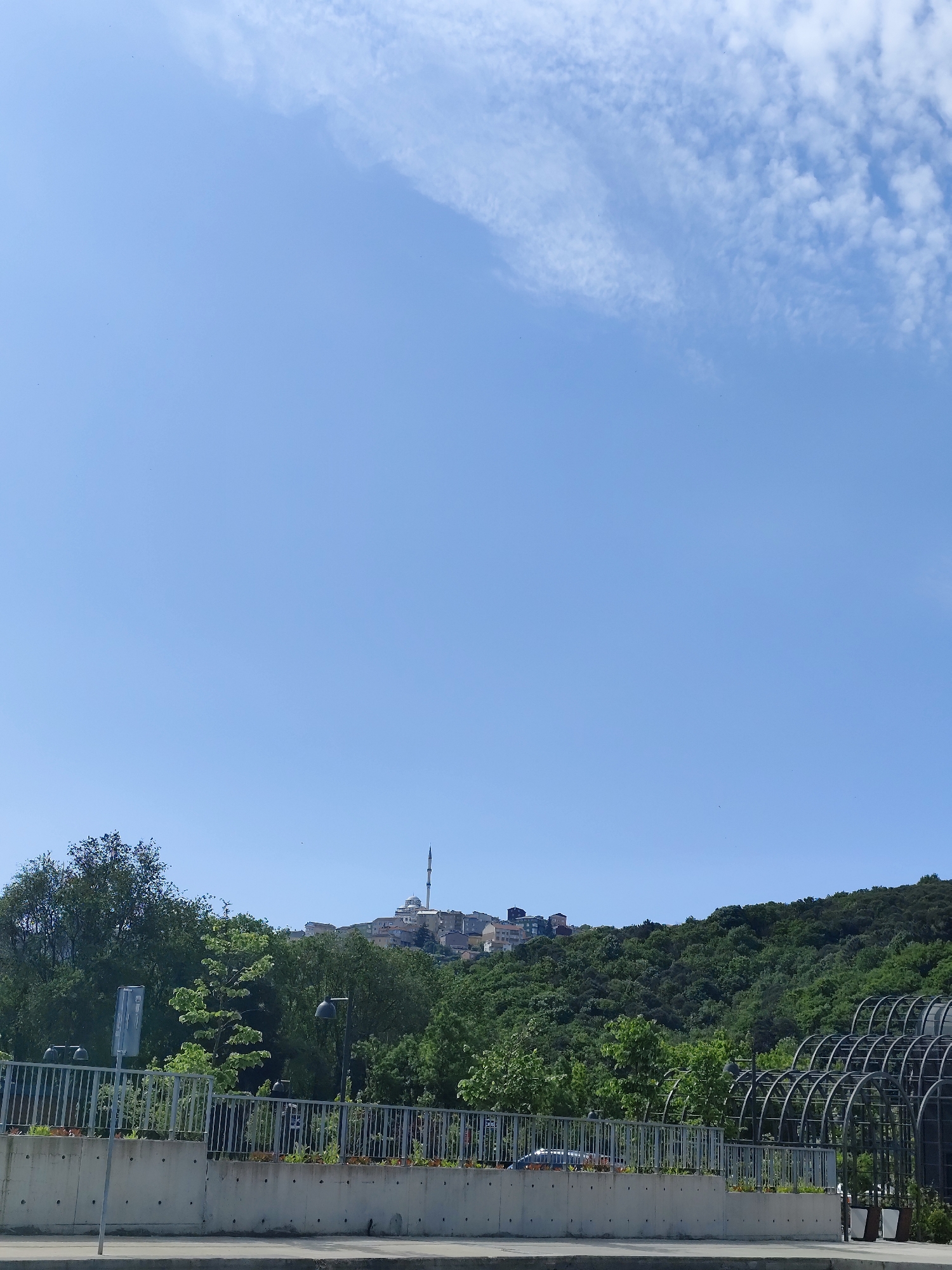 The image depicts a cityscape in Sarıyer, Turkey, under a clear, bright blue sky dotted with a few wispy white clouds. A mosque with a tall minaret is visible atop a hill, surrounded by residential buildings. Lush green trees cover the hillside, cascading down towards a concrete retaining wall with a metallic railing. Flowers are planted along the retaining wall. A metallic arched structure stands on the right side of the image. A road is visible in the foreground.