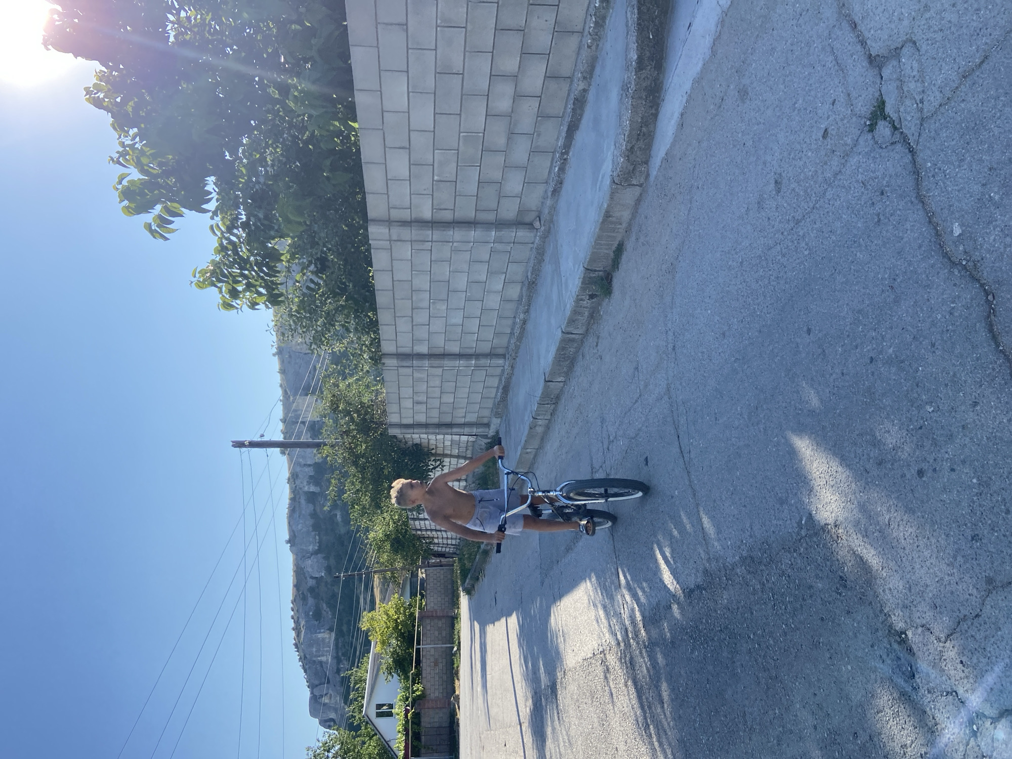 A shirtless boy with blonde hair, wearing blue shorts and sandals, rides a BMX bicycle down a paved street in Красномакское сельское поселение, Ukraine. The boy is facing to the left of the frame, and his body is turned slightly towards the camera. The street is lined with a concrete sidewalk and a tall, grey cinder block wall on the right side of the image. Behind the wall, trees and foliage are visible. In the background, a house with a white facade and a fence can be seen, as well as a large, rocky hillside under a clear blue sky. Power lines stretch across the sky, and the sun casts long shadows on the road, indicating it is late afternoon.