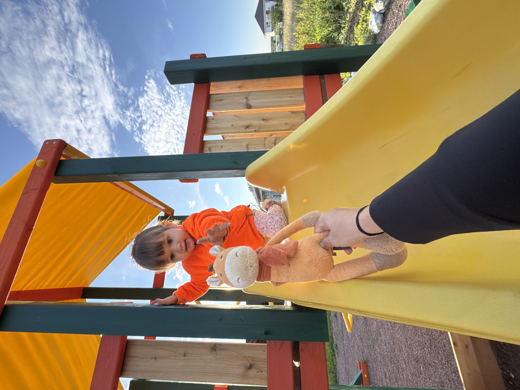 In Kempelevo, Russia, a young child with light brown hair, wearing an orange hooded jacket and patterned pants, is on a playground slide. The child is positioned on the upper part of a bright yellow plastic slide, with their left hand gripping a wooden support post. Their right arm is extended forward, reaching towards a stuffed giraffe toy. The toy giraffe, with a red scarf around its neck, is held by a person's hand visible in the foreground, with a black sleeve. The playground structure features orange and red accents, with wooden planks forming a wall behind the child. The background shows a clear blue sky with scattered white clouds, green grass, and houses in the distance. The ground beneath the slide appears to be covered in a reddish-brown material.