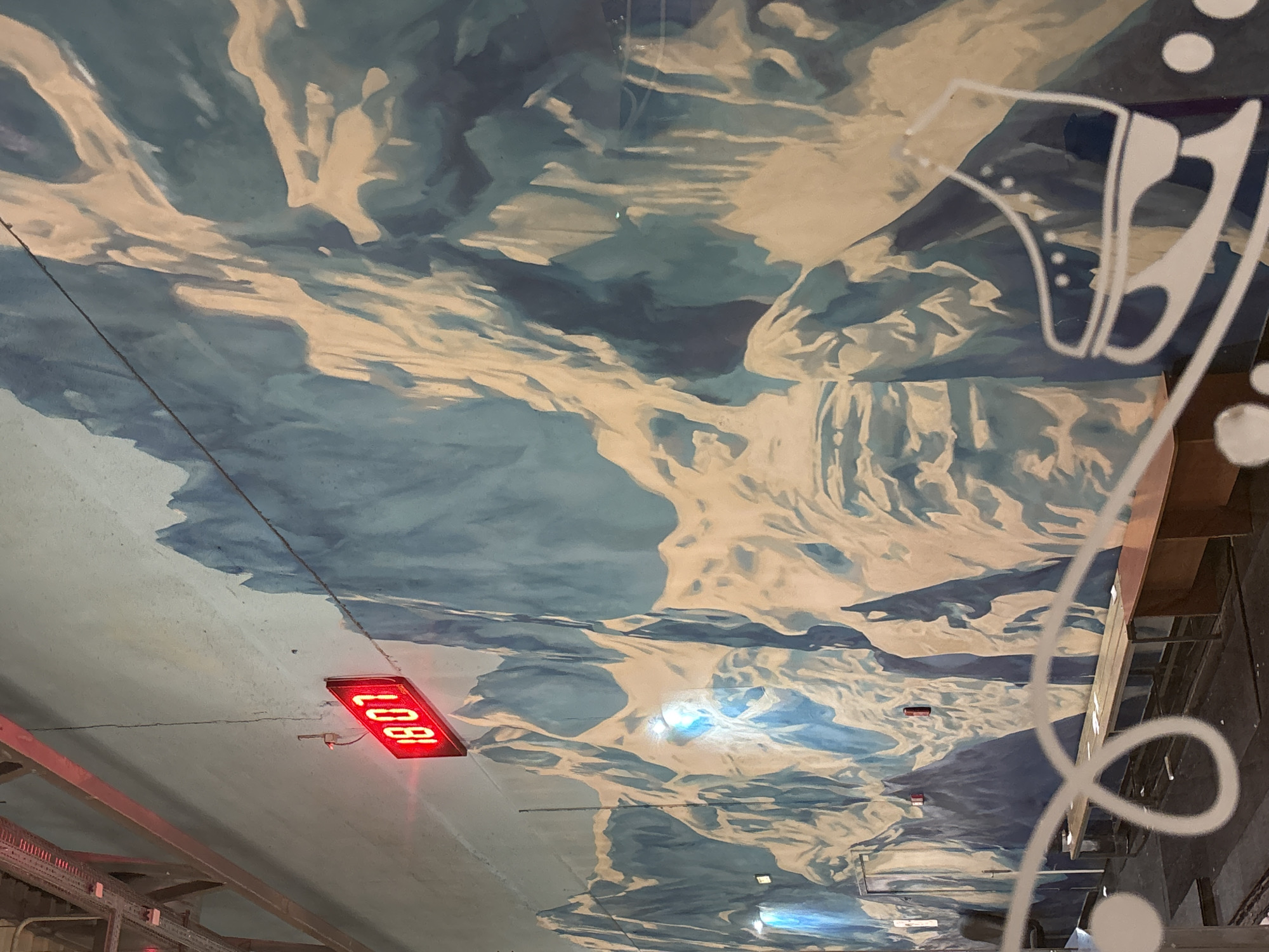 The image depicts the interior ceiling of the Minsk Airport National, located in Minsk, Belarus. The ceiling features a painted mural of mountains and sky in shades of blue, white, and beige. A rectangular LED sign displaying "1807" in red is mounted to the ceiling. A white, stylized design with circular and linear elements is visible on a transparent surface to the right of the image, partially obscuring the ceiling mural. Metal framework and mechanical elements are visible in the lower-left corner, suggestive of the airport's infrastructure. The overall perspective is from below, looking upward toward the ceiling.