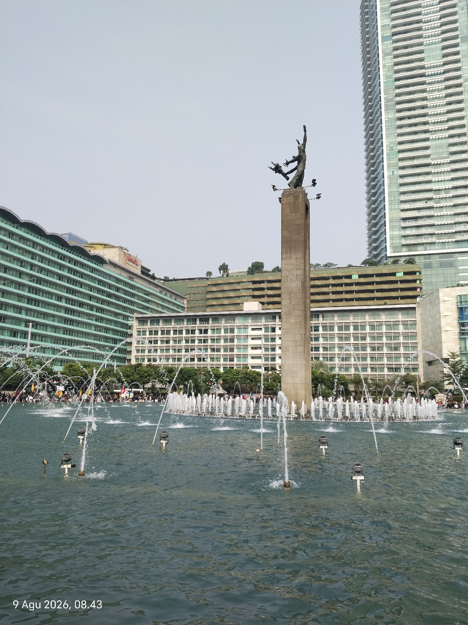 This image depicts the Arjuna Wiwaha Monument, a significant landmark located in Central Jakarta, Indonesia. The monument, a towering concrete pillar topped by a bronze sculpture of Arjuna and his celestial charioteer, Krishna, stands prominently in the foreground. The sculpture depicts Arjuna drawing his bow, signifying his victory in the Mahabharata epic.
In front of the monument, a large fountain sprays water, creating a dynamic and lively atmosphere. The water reflects the surrounding buildings, adding depth and visual interest to the scene. The fountain is surrounded by a body of water, with multiple water jets creating a curtain of spray.
Behind the monument, several buildings of varying architectural styles line the cityscape. To the left, a large, multi-story building with a green facade and arched windows dominates the view. To the right, a modern skyscraper with a glass facade and numerous horizontal windows pierces the sky. In between these structures, other buildings with white and beige facades are visible.
In the background, numerous people are gathered around the fountain area, suggesting it is a public space or a tourist attraction. Their presence adds a sense of scale and activity to the scene. The sky above is a clear, pale blue, indicating a bright and possibly sunny day. The overall impression is of a bustling urban environment with a blend of historical and modern architecture, centered around the iconic monument and its accompanying fountain.