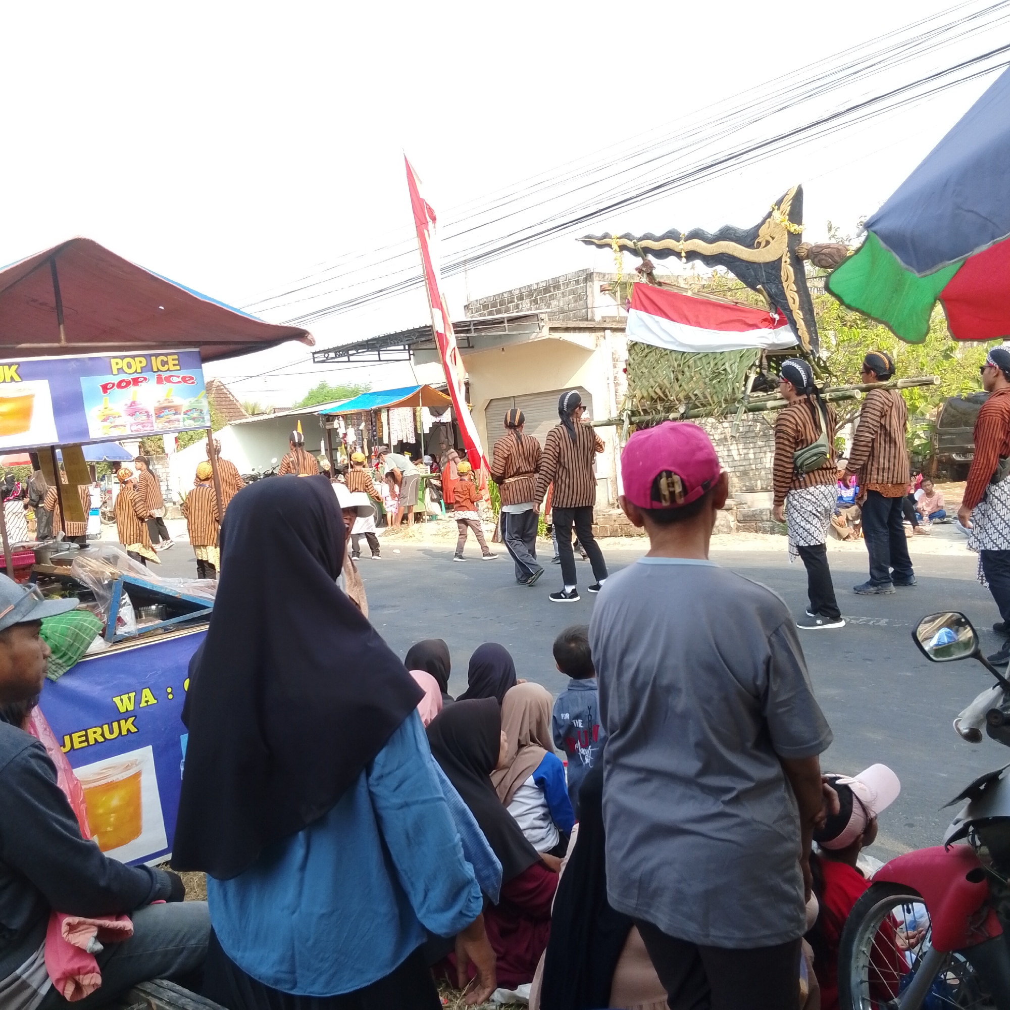 The image depicts a street scene in Plumpang, Indonesia, likely during a cultural event or parade. In the foreground, several individuals are visible, some wearing traditional attire. A person in a black hijab and blue shirt faces away from the camera, while another in a grey shirt is positioned to the left, also looking away. A child wearing a pink cap is in the center foreground.
In the middle ground, several people dressed in striped brown and white traditional outfits are walking or standing. Some are carrying bamboo poles over their shoulders. The Indonesian flag, red and white, is prominently displayed, along with a decorative keris (dagger) on a stand. There are also food stalls or vendors visible, one with a sign displaying "POP ICE."
The background shows buildings and a street with overhead power lines. Several motorcycles are parked, suggesting a public gathering. The overall atmosphere appears to be festive and community-oriented. The lighting suggests it is daytime, with bright sunlight casting shadows.