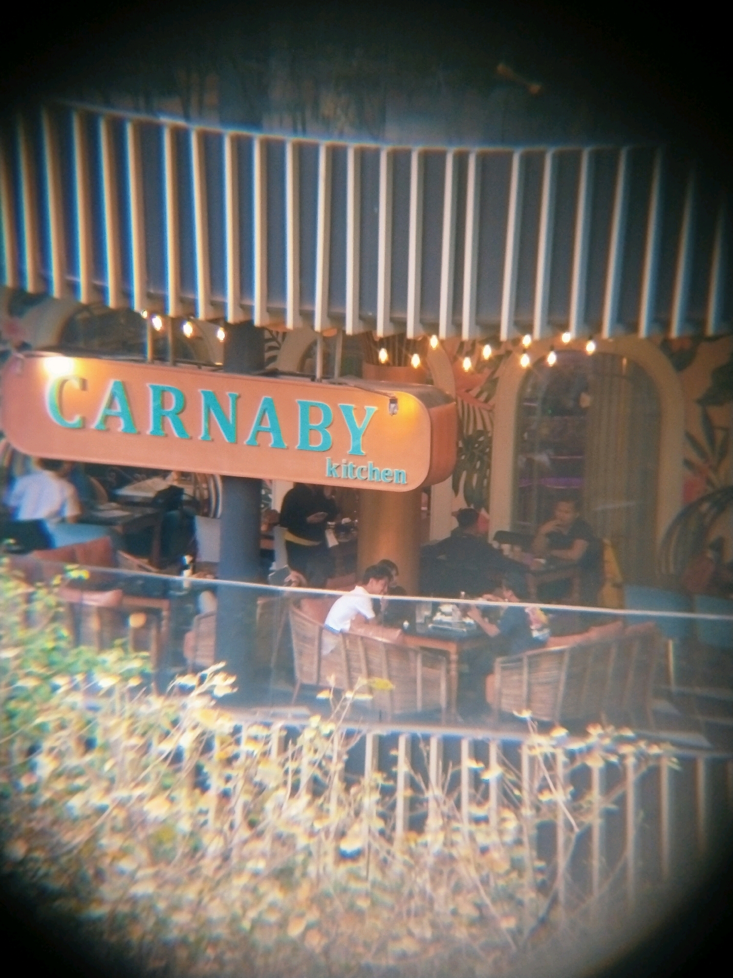The image depicts an outdoor seating area of a restaurant, likely in Surabaya, Indonesia, with a sign that reads "CARNABY kitchen". The setting appears to be a casual dining establishment with patrons seated at tables. In the foreground, there is a blurred view of greenery, suggesting foliage or plants. The background shows the restaurant's architecture, including a slatted roof structure and a decorative wall with tropical-themed art. Several people are visible, some seated at tables, while others are standing or moving around. One person is clearly visible in the foreground on the right, wearing a white shirt and looking towards the restaurant. The lighting suggests it might be daytime or early evening. The overall impression is that of a lively, open-air dining environment.