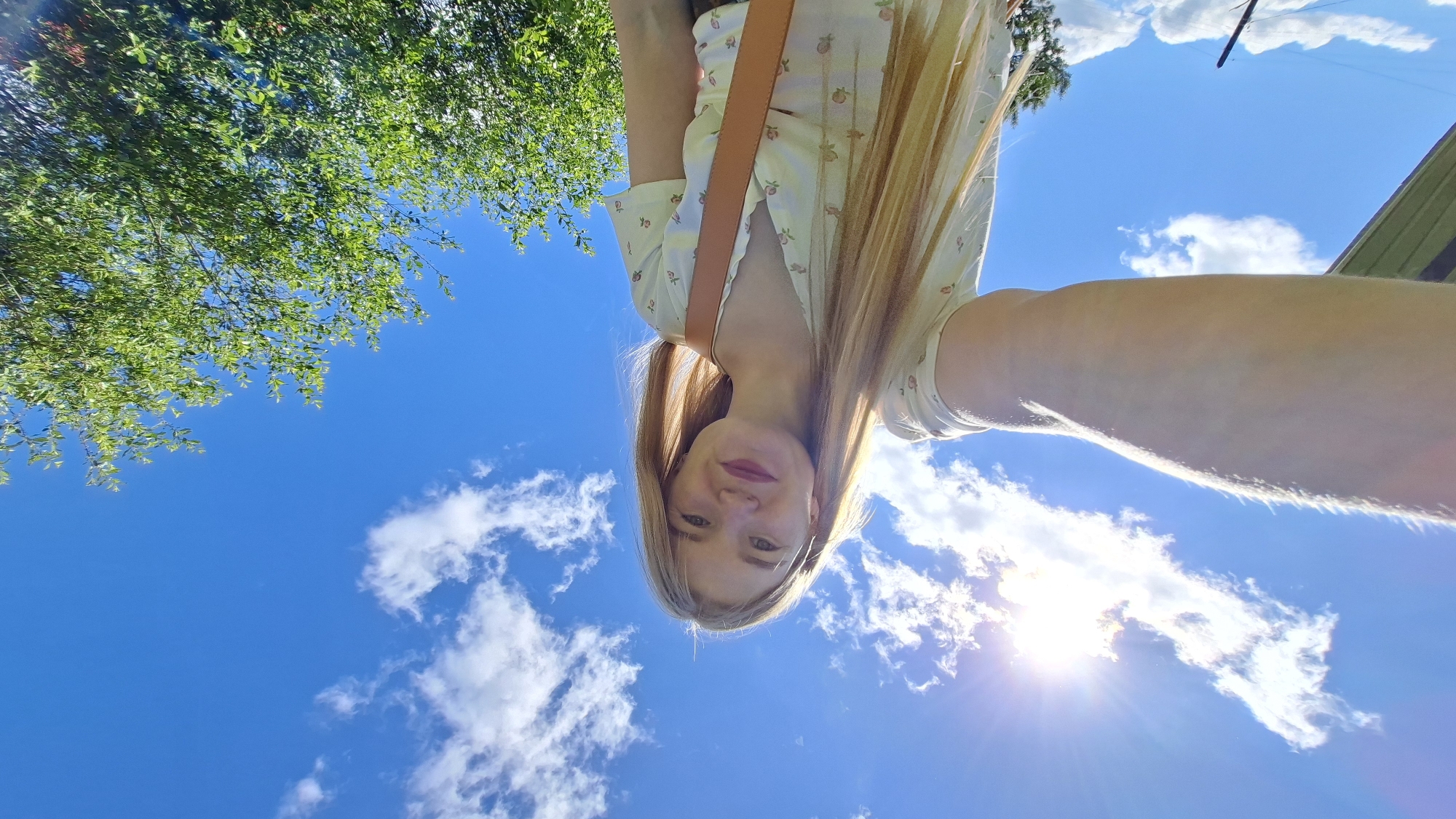 A young woman with long blonde hair is taking a selfie outdoors under a bright blue sky with scattered white clouds. She is wearing a light-colored dress with a floral pattern and a brown strap across her chest. The sun is shining intensely, causing lens flare in the upper left corner of the image. To the right, a lush green tree is visible, with branches extending into the frame. In the background, a thin, dark vertical pole or stick is present, and a glimpse of what appears to be a green structure, possibly a building or fence, can be seen on the far left. The perspective is low-angle, looking up at the woman and the sky. The scene is set in the village of Bolshoy Bukor, Russia, indicated by the context provided.