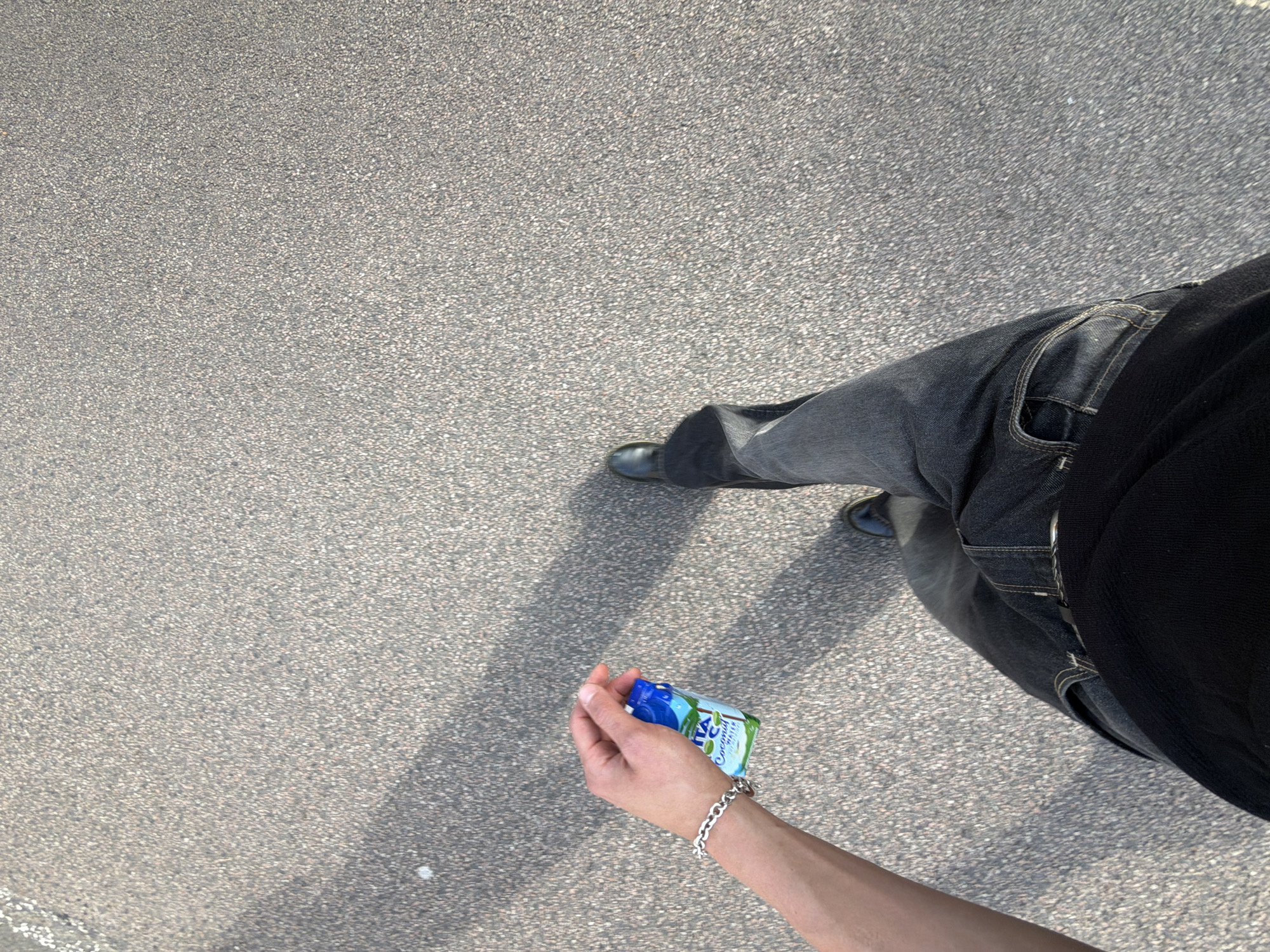 A first-person perspective shot shows a person walking on a paved surface. The person is wearing dark-wash jeans, black boots, and a black t-shirt. They are holding a small, rectangular blue and white juice box in their right hand. A silver bracelet is visible on their left wrist. The paved surface appears to be asphalt with small, light-colored pebbles. Shadows of unseen objects fall across the ground, indicating daylight. No specific London landmarks or street signs are visible, making it impossible to determine the precise location within London. The overall scene is ordinary, depicting a person in transit on a street or path.