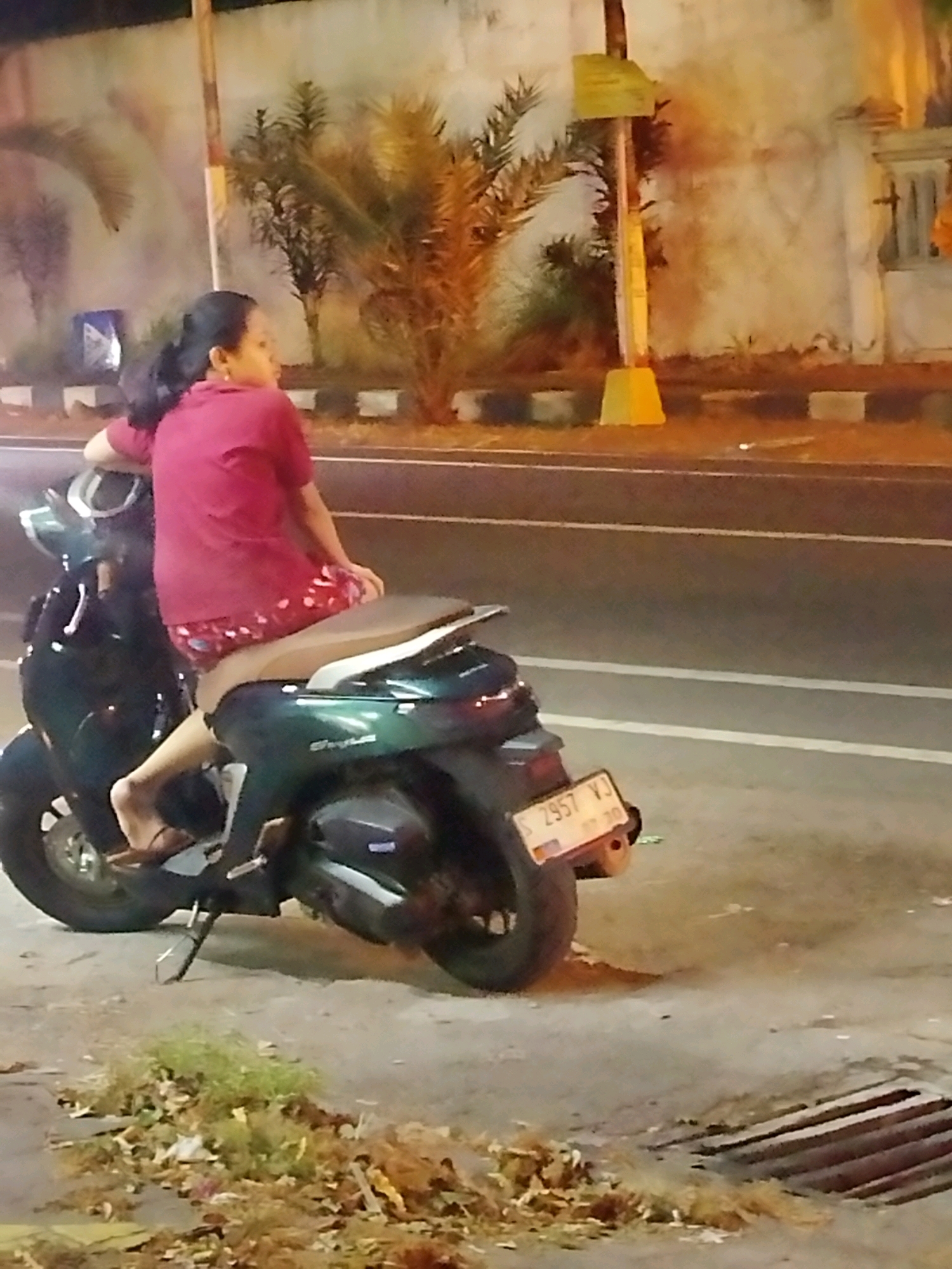 A woman with dark hair pulled back is sitting on a dark green scooter parked on the side of a road at night. She is wearing a reddish-pink short-sleeved shirt and patterned shorts. Her left leg is resting on the footrest of the scooter, and her right foot is on the ground. The scooter has a license plate that reads "S 2957 YJ". In the background, there is a wall, a street light, and some palm trees. The ground is paved with concrete, and there is some debris scattered around. The scene appears to be in a city in Mojokerto, Indonesia, based on the license plate format.
