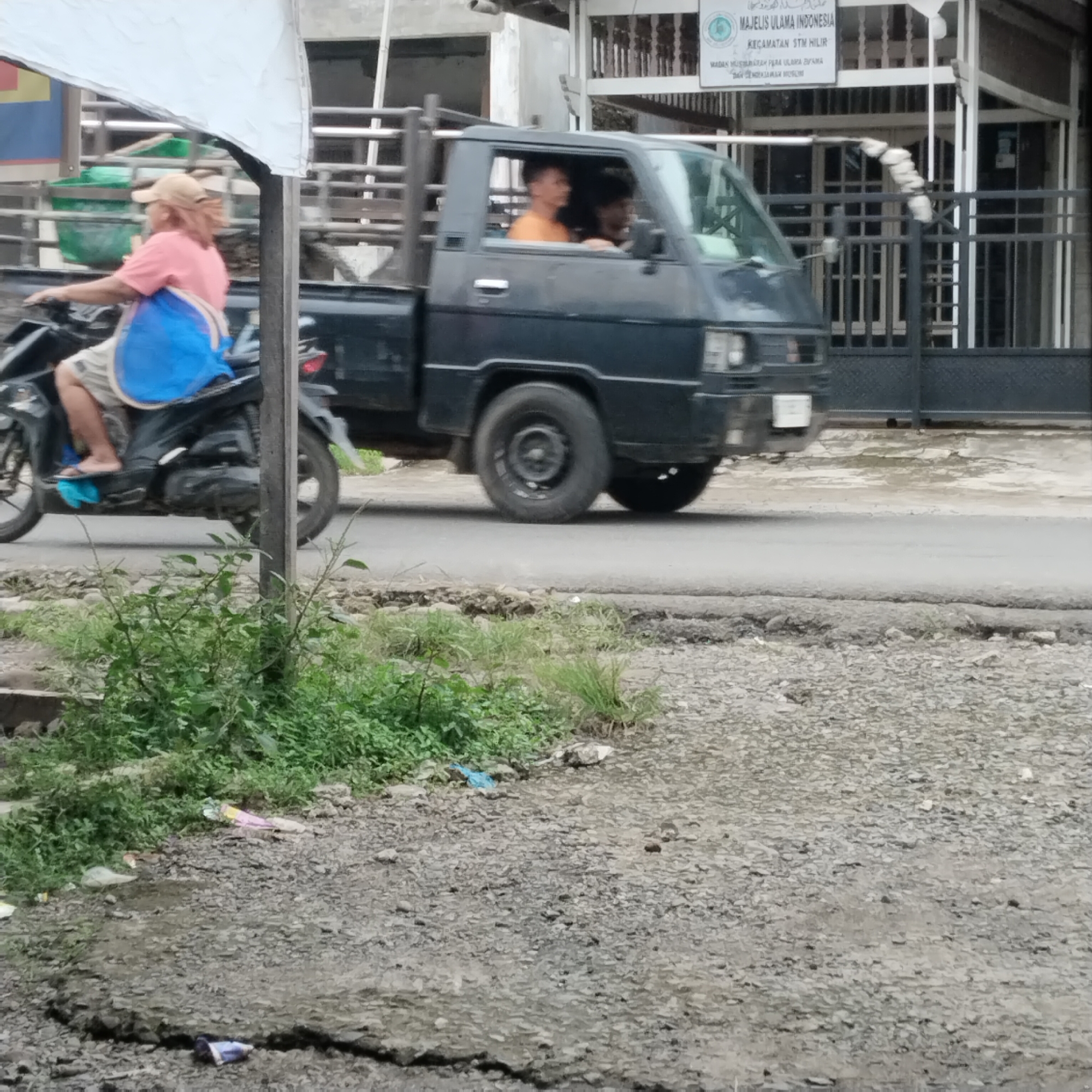 In Talun Kenas, Indonesia, a man wearing a pink t-shirt and a beige cap rides a motorcycle on a paved road. He is carrying a blue bag on his back. To his right, a dark-colored pickup truck is driving, with two men inside the cab. The driver, wearing an orange shirt, is visible. The truck has a metal cage on the back, and a white tarp is draped over a structure to the left of the truck, partially obscuring a yellow and blue sign. The roadside is covered in gravel and sparse green vegetation, with some litter visible on the ground, including a blue plastic bag. In the background, a building with white pillars and a dark metal fence can be seen, with a sign on the building that reads "MAJELIS ULAMA INDONESIA" and "KECAMATAN".