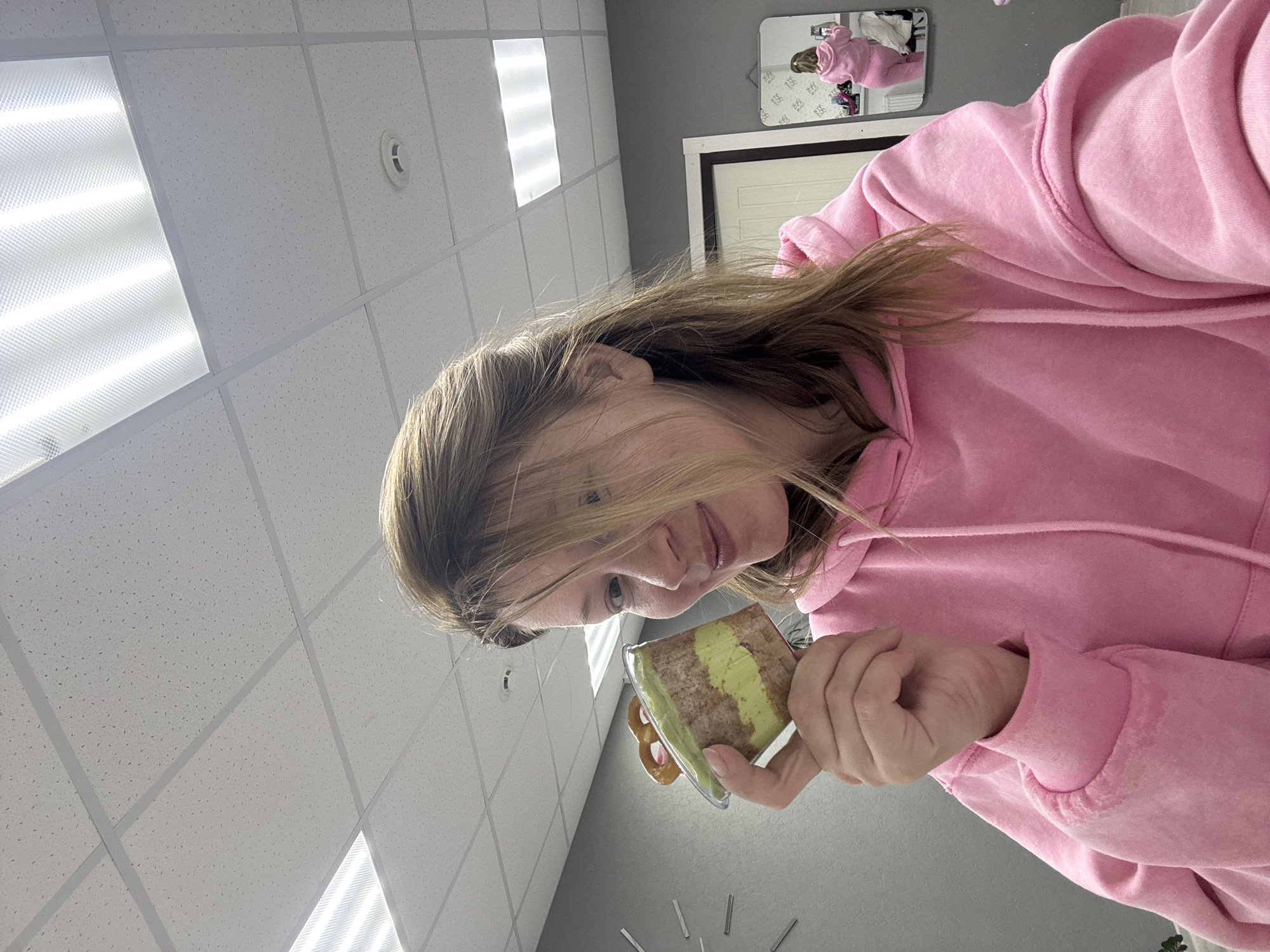 An indoor, vertically oriented close-up shot from Chaikovsky, Russia, features a young woman holding a layered dessert. The woman, with long, light brown hair partially obscuring her face, is wearing a bright pink hooded sweatshirt and looks down at the dessert with a subtle smile. Her right hand, with manicured nails, holds the clear plastic container.
The dessert shows distinct layers, including a prominent greenish layer (possibly matcha or pistachio) and a brownish cake-like layer, with a pretzel stick also visible within the container.
The background reveals a typical office or commercial interior with a white acoustic tile drop ceiling, brightly illuminated by rectangular fluorescent light fixtures. One fixture, casting a strong light, dominates the upper left. A circular white vent is also seen on the ceiling. The walls are a neutral grey. A small, decorative mirror on the wall reflects a figure in a pink top, suggesting either a self-portrait setup or another person. Part of a white door with a dark frame is visible next to the mirror. Below the woman's hand, a modern wall decoration resembling a clock face with silver markers is partially visible. The lighting is artificial and bright. No discernible text is present in the image. The overall impression is one of casual contentment.
