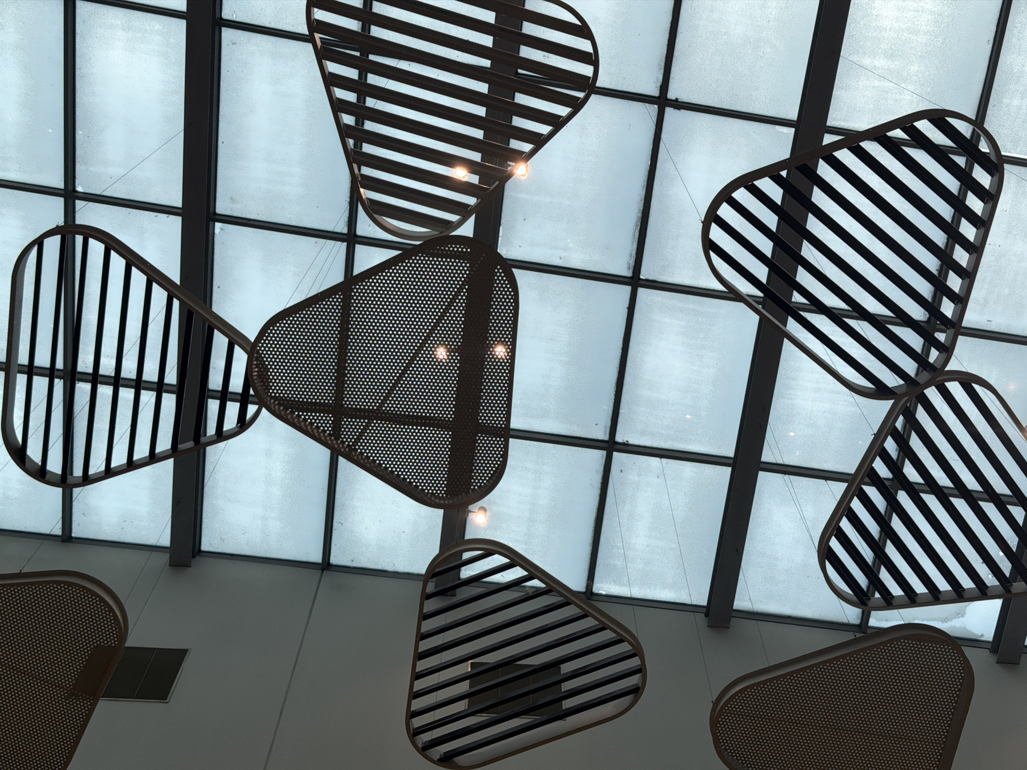 The image displays an upward view of a contemporary interior ceiling, characterized by a large, grid-patterned glass skylight and several artistic light fixtures suspended beneath it. The skylight is composed of rectangular glass panes separated by dark, possibly metal, framing. The glass surfaces appear diffused, textured, or covered with a light layer of frost or condensation, suggesting overcast conditions or cold weather outside, which filters the natural light entering the space.
Hanging from the ceiling are multiple abstract, triangular or teardrop-shaped objects. These serve as both decorative elements and luminaires. Some of these fixtures feature a slatted design, with dark, thin strips arranged diagonally or vertically within a curved, light-colored frame. Other fixtures are made from a darker material with a fine, repetitive perforated pattern, resembling a mesh or cane work. Small, exposed light bulbs emit a warm glow from within or behind some of these fixtures, contrasting with the cool, diffused light from the skylight.
The lower portion of the image shows a plain, light-colored ceiling or wall area. There are no people visible, and no specific activity is taking place; the focus is on the architectural and design elements. The aesthetic is modern and clean, creating a calm atmosphere. Given the context of Sofia, Bulgaria, this scene likely captures an interior space such as a shopping mall, public building, or a large commercial establishment, showcasing its unique ceiling design and lighting arrangements. No visible text is present in the image.