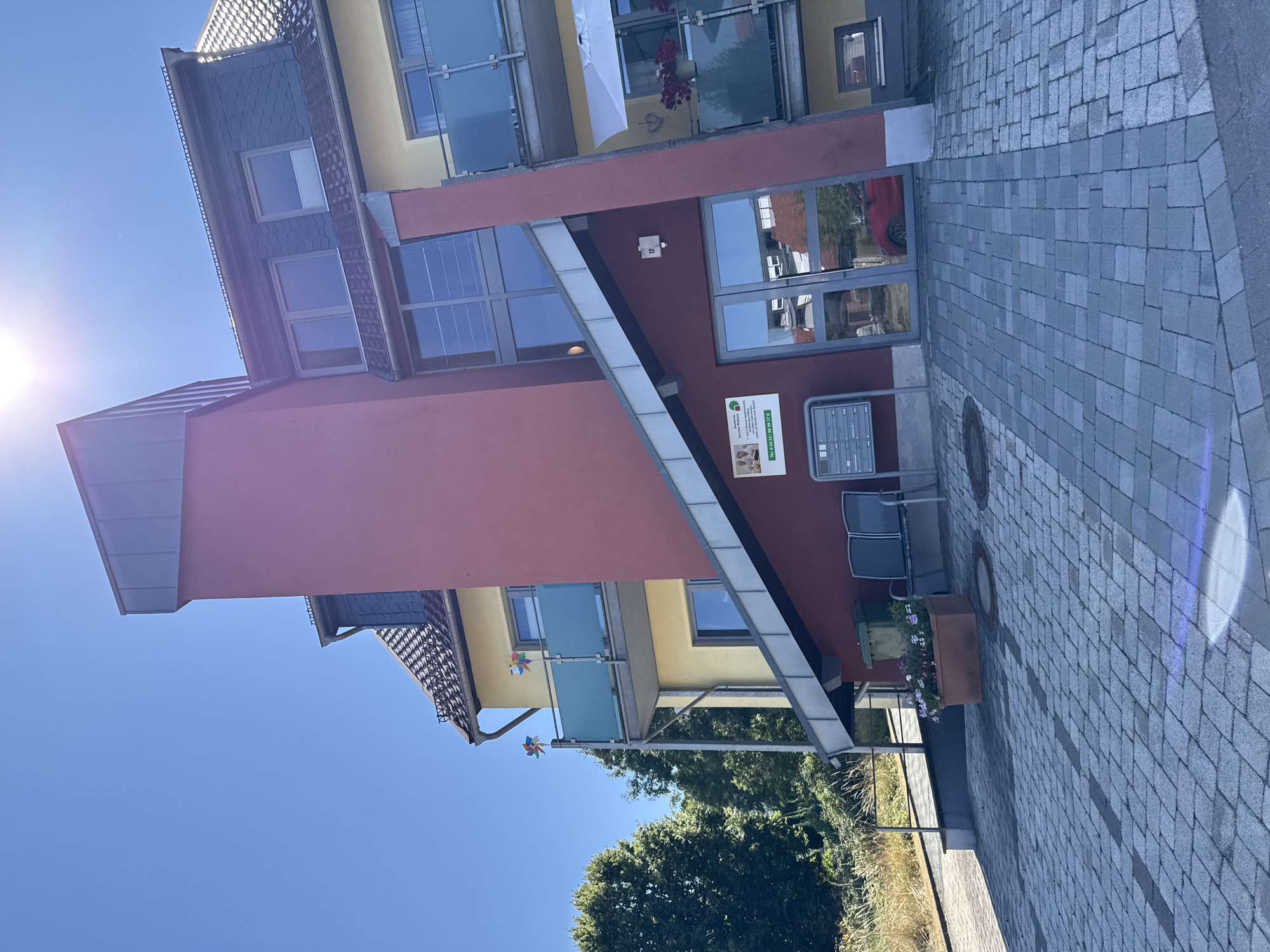 The image shows the exterior of a residential building in Bad Sachsa, Germany, on a clear, sunny day. The building features a striking architectural design with contrasting colors, primarily a deep red on the main facade and a pale yellow on the upper stories. It has multiple levels with balconies, some of which have green glass railings. There are several windows visible, including large floor-to-ceiling windows on one side. A modern, angled roofline with dark grey shingles and metal elements is also prominent.
At the ground level, there is a paved walkway made of grey interlocking bricks. In front of the building, a mailbox unit is mounted on the red wall. To the left of the entrance, there is a sign with German text and images, likely indicating a business or community service. Adjacent to the sign are two dark grey benches and a green trash bin. A large rectangular planter box filled with flowers sits on the paving stones.
The entrance itself appears to be double glass doors with a metal frame. A red car is partially visible reflected in the glass doors. To the left of the building, a lush green tree line forms a backdrop, and a metal railing leads up a sloped pathway. Two colorful pinwheels are mounted on the railing of an upper balcony. The sky is a vibrant blue with a bright sun, creating lens flare in the upper right portion of the image. The overall impression is of a modern, well-maintained building in a suburban or semi-urban setting.