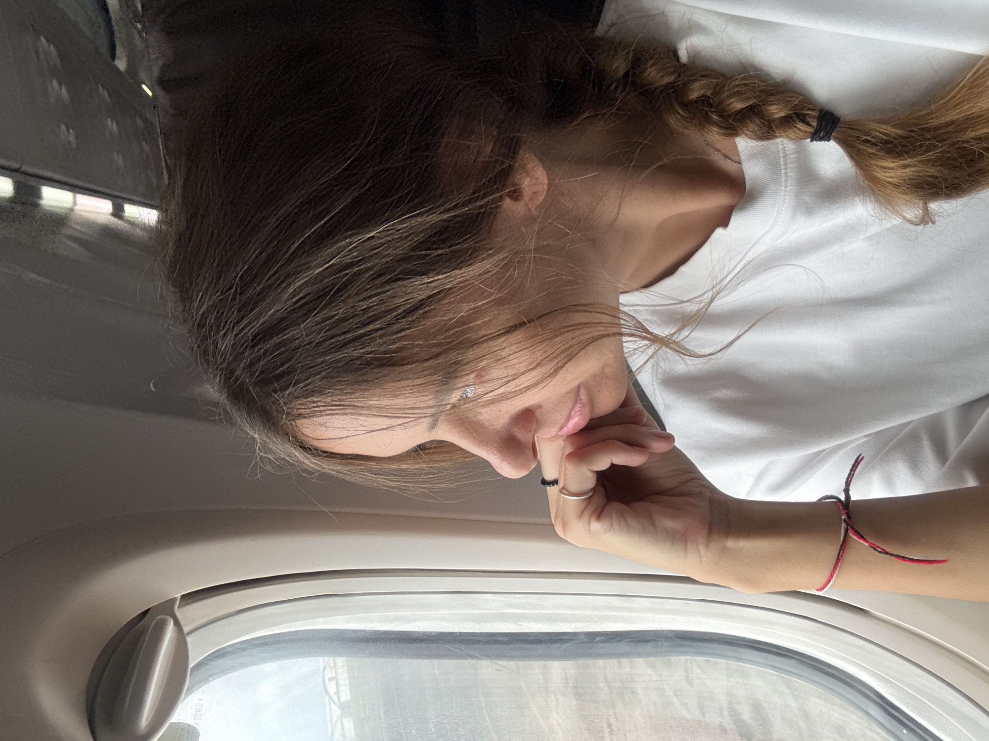 A woman with braided hair, fair skin, and a white t-shirt is pictured from a slightly above angle, looking out an airplane window. She is resting her chin on her hand, and has several rings on her fingers. A red and white string bracelet adorns her wrist. The view outside the window shows elements that could be buildings or structures. The location is specified as Kuta, Indonesia, which implies the woman is on a plane arriving at or departing from the area.