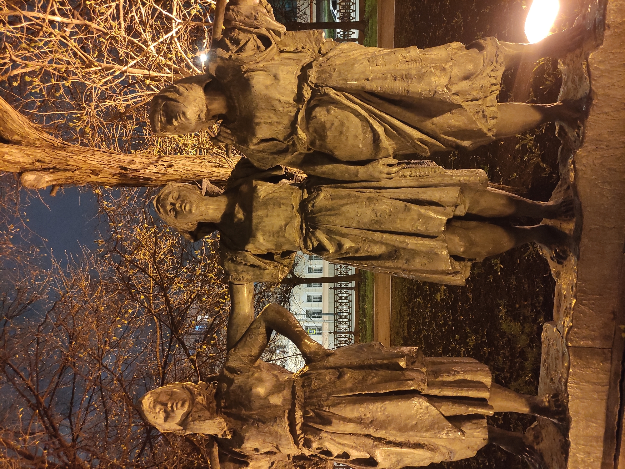 The image, taken at night, features three bronze-colored statues of women positioned in an outdoor setting, illuminated by warm artificial light. The statues stand upright, depicting female figures in long, draped garments with a stylized, textured finish.
The figure on the left appears to be leaning slightly forward, her head tilted, possibly engaged with an unseen element or holding an object. The central figure stands with her arms seemingly occupied in front of her, perhaps carrying a bundle or interacting with another element. The third figure, on the right, stands tall and appears to be looking upwards, with one arm slightly raised. Their facial expressions are subdued, suggesting a narrative or allegorical theme.
Behind the statues, a prominent bare tree with numerous branches, possibly in late autumn or early spring, is brightly lit, casting a warm glow. The night sky above is a deep blue. In the background, a multi-story building with illuminated windows is visible through the tree branches, indicating an urban environment. Greenery, likely bushes or low plants, can be seen at the base of the statues. A bright light source, possibly a streetlamp, is prominent on the far right, casting intense light. There is no visible text in the image. The scene depicts a solemn and reflective monument in an urban area at night, consistent with a public space in Moscow, Russia.