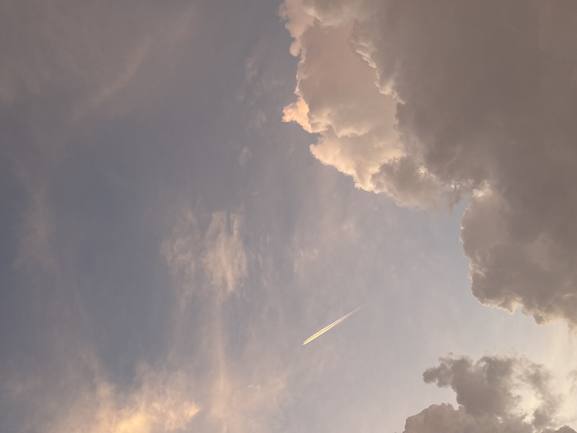 The image displays an expansive view of a partially cloudy sky, rendered in soft, ethereal light suggestive of late afternoon or early evening. The sky transitions from a gentle blue-grey on the left to warmer, golden, and pinkish hues on the right, indicating sunlight catching the clouds at a low angle.
Large, voluminous clouds dominate the upper right and lower right portions of the frame. These clouds exhibit significant depth and texture, with their edges brightly illuminated by warm, golden-pink light, while their interior masses are cast in darker shades of grey and beige, creating dramatic contrast. Scattered across the mid-left and upper-left sections are thinner, more diffused wisps of clouds, blending smoothly into the sky.
A prominent feature is a long, luminous contrail (vapor trail) streaking diagonally across the sky from the lower-left to the upper-right quadrant. The contrail itself glows with a subtle yellowish-white hue, reflecting the golden light. At the leading edge of this trail, a small, dark silhouette of an aircraft is visible, indicating its high altitude.
The overall scene conveys a sense of peaceful vastness and natural beauty, with the interplay of light and shadow on the clouds creating a serene atmosphere. The image captures a moment of tranquil flight against a backdrop of dynamic sky patterns, observed from Choeng Thale, Thailand. No text is visible in the media.