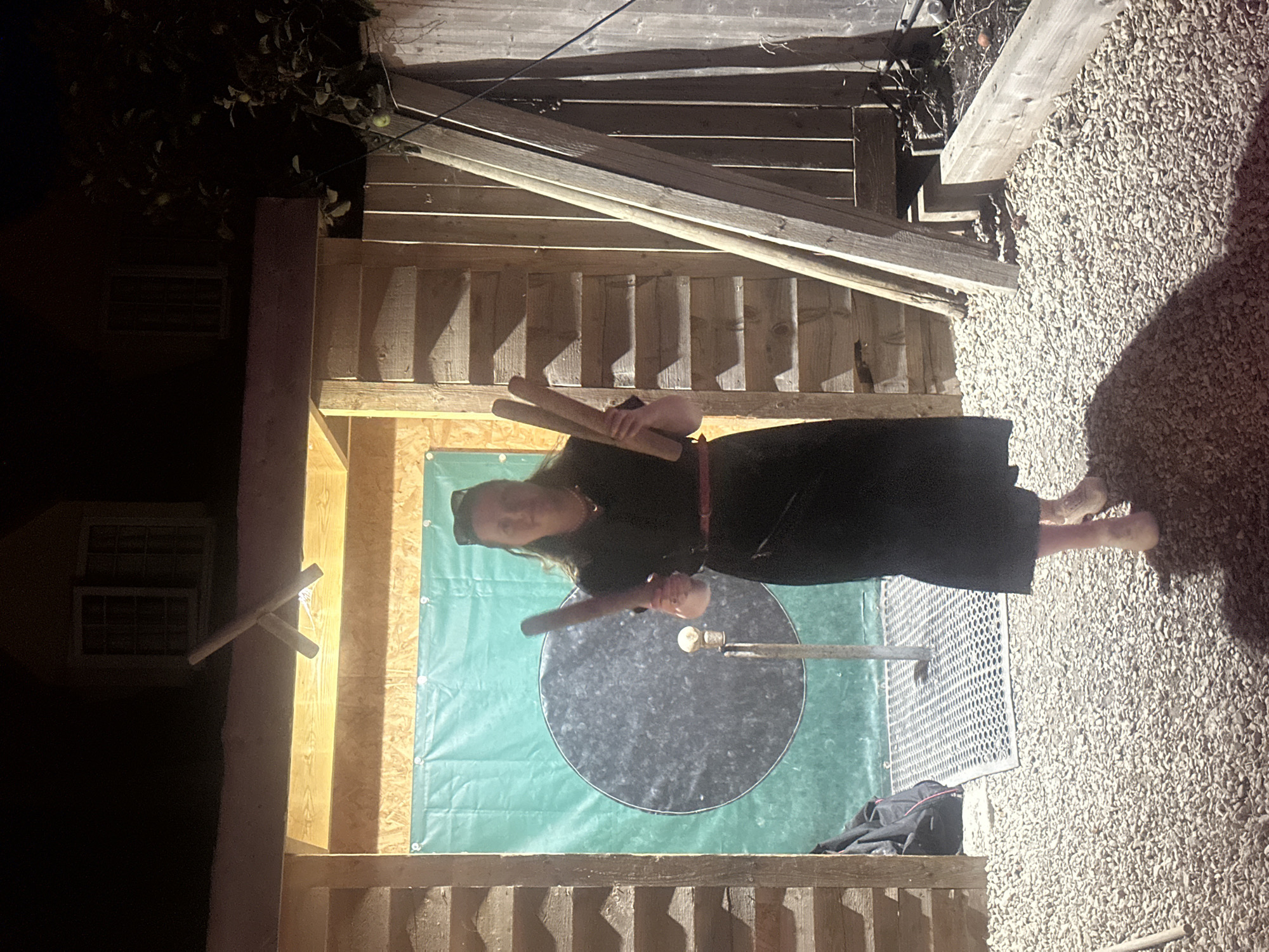 A woman stands in a gravel-covered outdoor area at night. She is wearing a black dress with a red belt and light-colored shoes. She holds three wooden mallets. Behind her is a structure made of wood with a green tarp hanging on the back wall, on which a large black circle is visible. The structure is illuminated by artificial light. A building with windows is visible in the background above the structure.