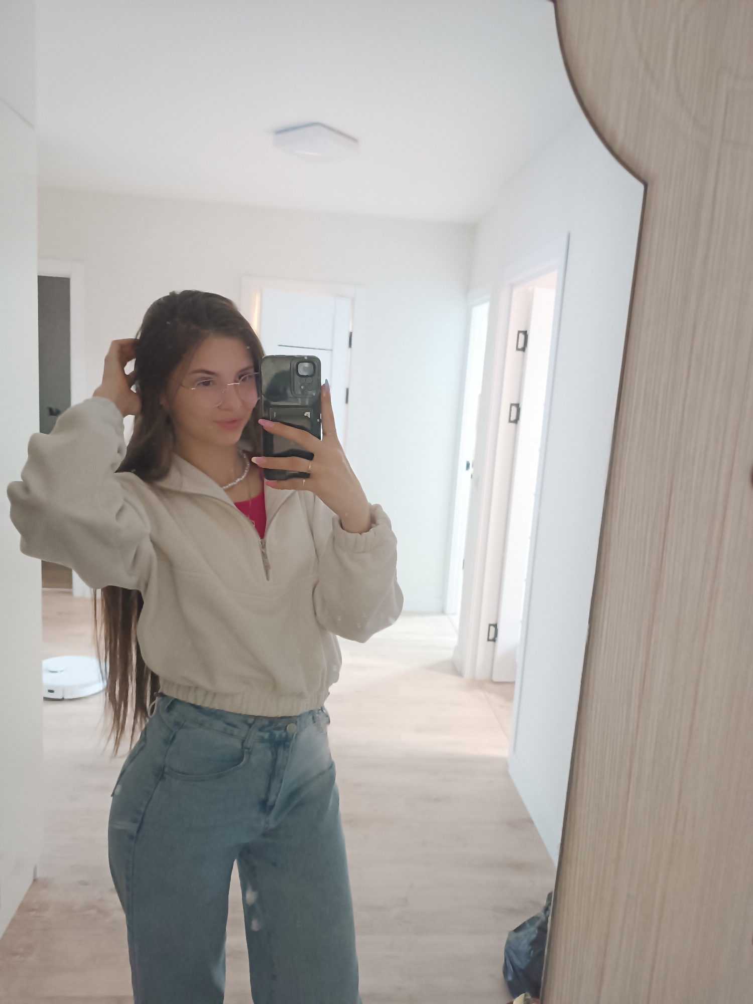 A woman with long brown hair, wearing wire-rimmed glasses, a cream-colored pullover, a pink shirt, jeans, and a pearl necklace, is taking a selfie in a mirror. She is holding a black smartphone in her right hand and touching her hair with her left hand. The mirror is leaning against a wall in what appears to be a hallway. Behind her is a hallway with several doors and a robot vacuum cleaner on the floor. The location is Unknown, Unknown.