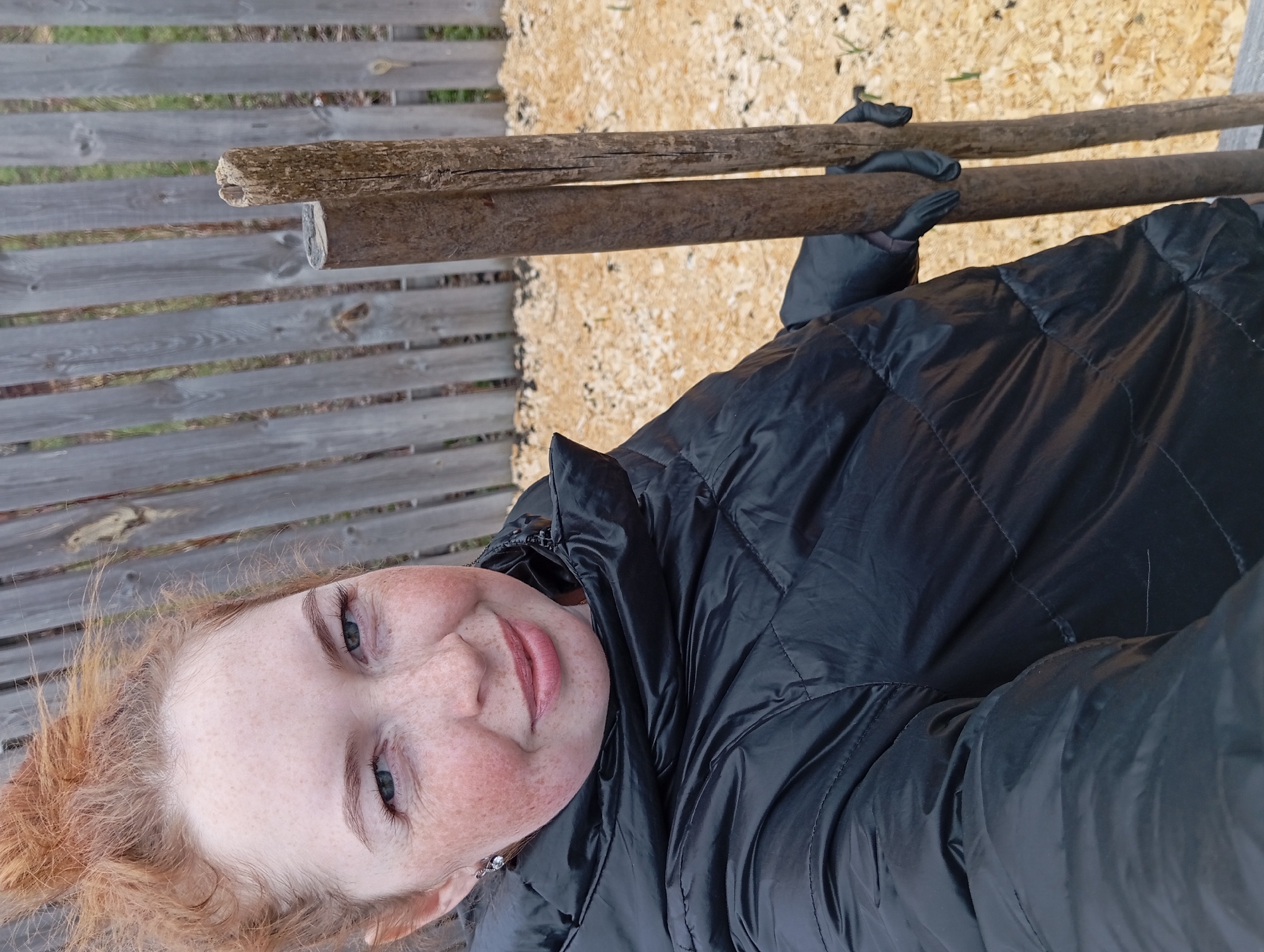 The image is a selfie taken by a fair-skinned woman with reddish hair and freckles. She is wearing a black puffy jacket and black gloves, and is holding two wooden posts. Behind her, there is a wooden fence on the left and wood chips on the right. The location is Yurgamysh, Russia.