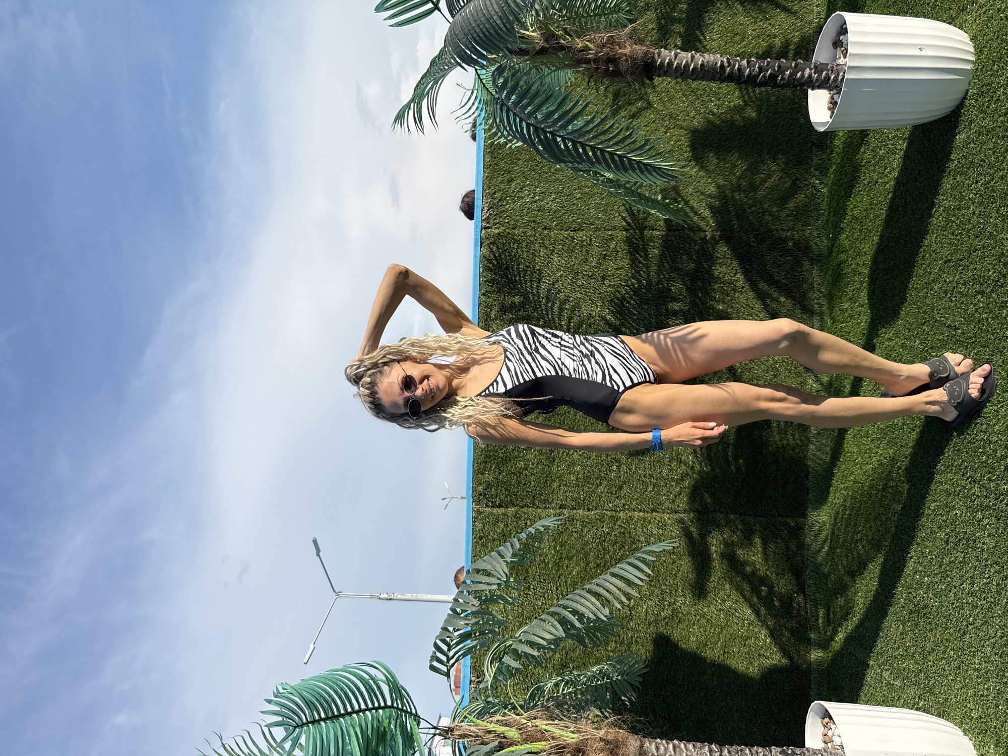 A woman wearing a black and white zebra-striped swimsuit is posing on an artificial grass surface in front of a blue and white structure, with artificial palm trees and a lamp post visible in the background. The location appears to be outdoors, possibly in Rusanovo, Russia. The woman is wearing sunglasses and has her hand raised near her head. There are shadows cast on the grass, and the sky is visible above.