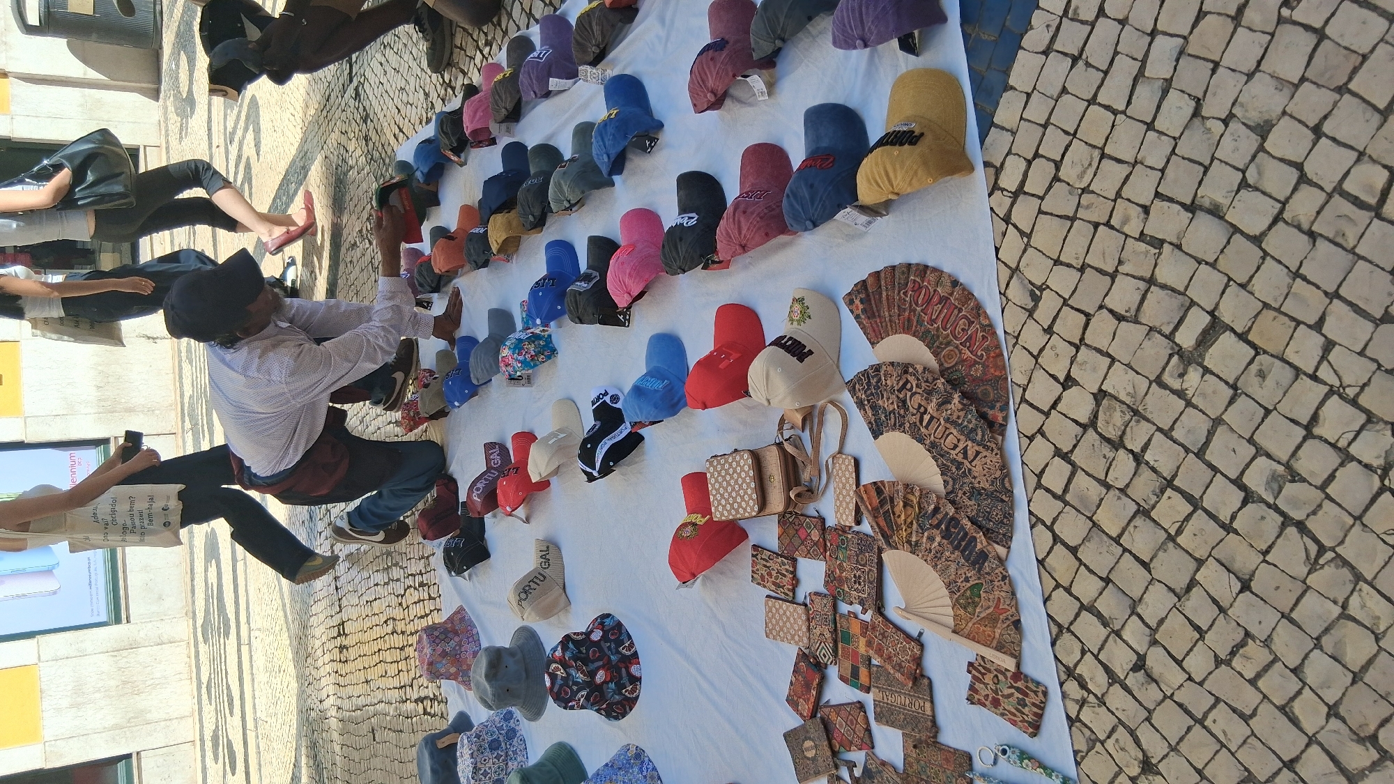 A street vendor in Lisbon, Portugal, is kneeling on the cobblestone pavement and arranging merchandise on a white tarp. The merchandise includes an assortment of baseball caps, bucket hats, and decorative fans, many of which feature the word "PORTUGAL" or other Portuguese motifs. A person in the background, partially visible, walks past, carrying a white tote bag. The scene appears to be set up outdoors, likely in a public area where tourists might frequent, given the nature of the goods displayed.