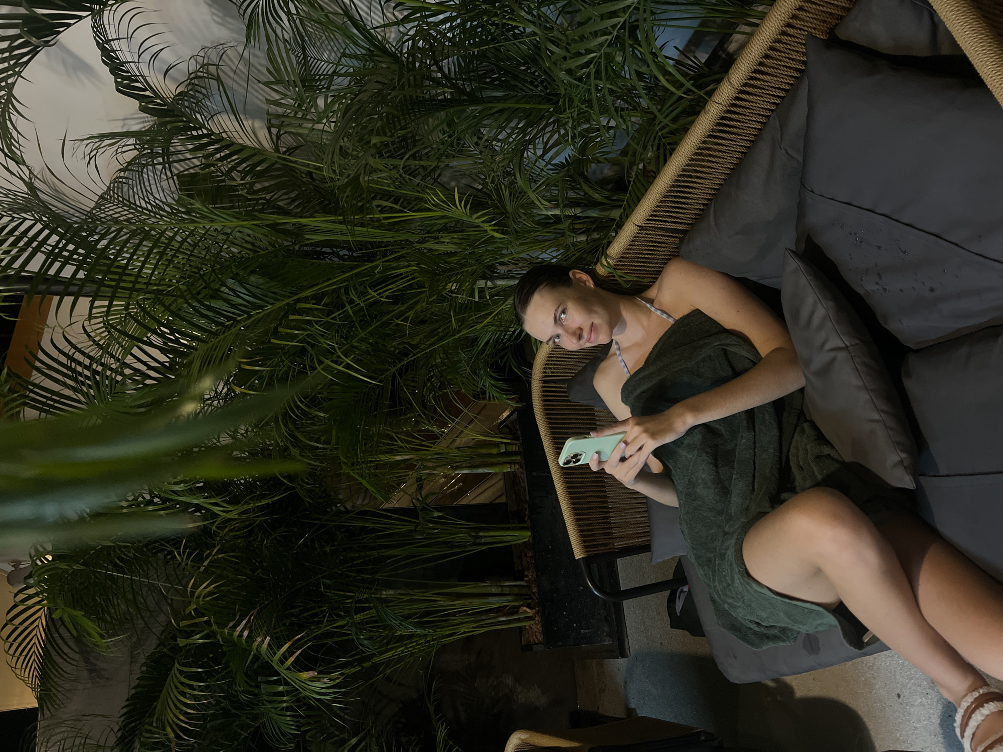 A woman is reclined on a gray cushioned patio sofa, wrapped in a green towel, and using a smartphone. The sofa is partially enclosed by a woven wicker-like structure. Lush green tropical plants surround the seating area, adding a natural, possibly private, ambiance. The location appears to be outdoors in what may be Si Sunthon, Thailand.