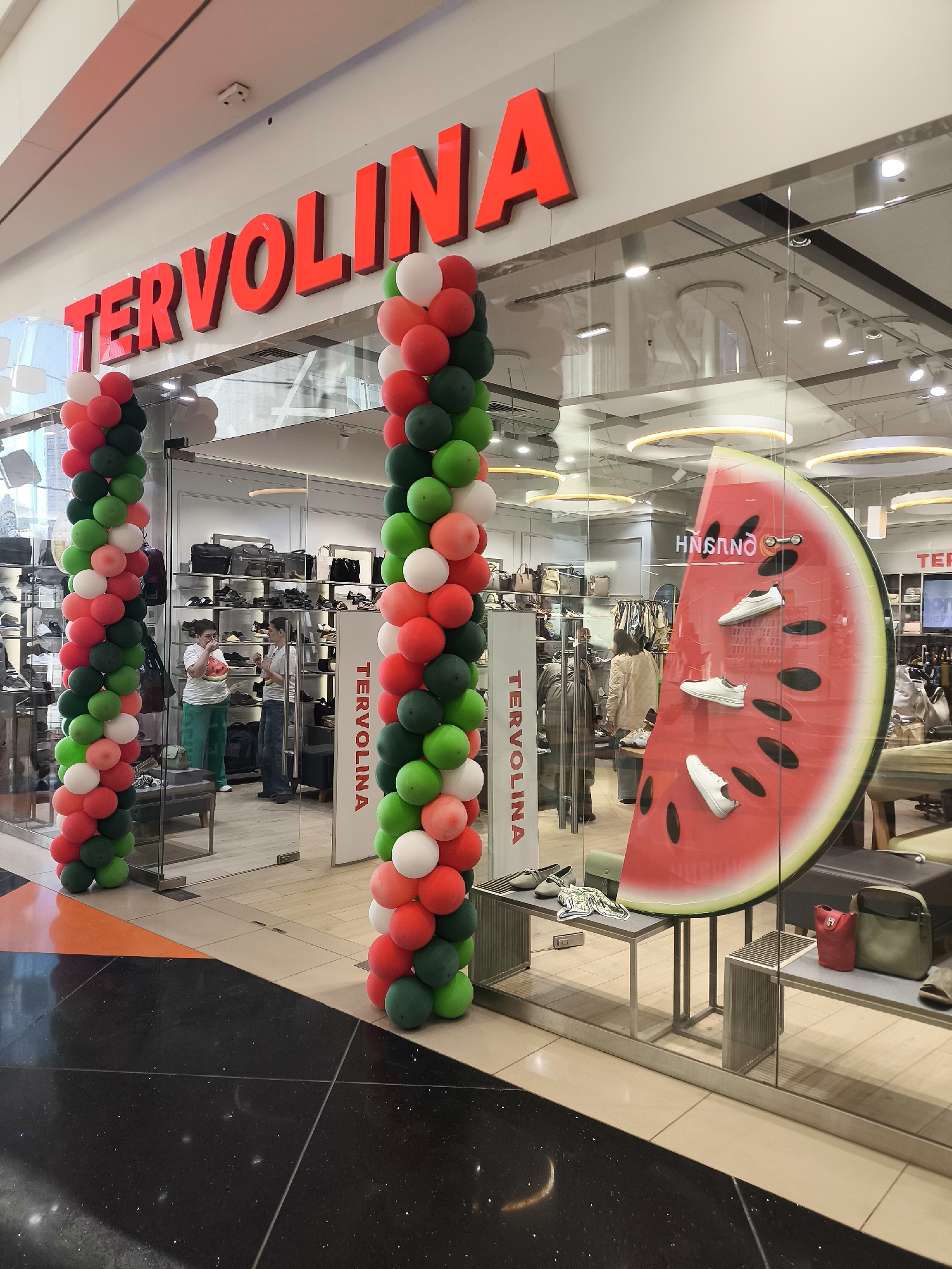 This image depicts the brightly decorated entrance of a retail store named "TERVOLINA," likely situated within a shopping mall in Khimki, Russia. The store's name is prominently displayed in large, red, illuminated letters above the entrance.
The storefront is festively adorned with two tall columns of red, white, and green balloons, suggesting a grand opening, special event, or celebration. The entrance consists of large glass panels, offering a clear view into the store's interior, which features display shelves filled with shoes and handbags under bright artificial lighting.
On the right side, the display window showcases a striking, oversized prop resembling a half-slice of watermelon, against which three white sneakers are mounted. Below this, various handbags in shades like grey, red, and olive green are displayed on modern metal shelving units. A vertical white sign with "TERVOLINA" written in red is visible on the right balloon column.
Inside the store, several individuals are present. On the left, through the glass, a person wearing green pants and a white top is visible, possibly interacting with an item, while another person browses merchandise further inside. On the right, another individual in a light-colored jacket is seen near display areas.
The floor in the foreground is a dark, sparkling tiled surface, transitioning to lighter beige and orange tiles directly in front of the store's entrance. The overall scene suggests a lively and inviting commercial environment.