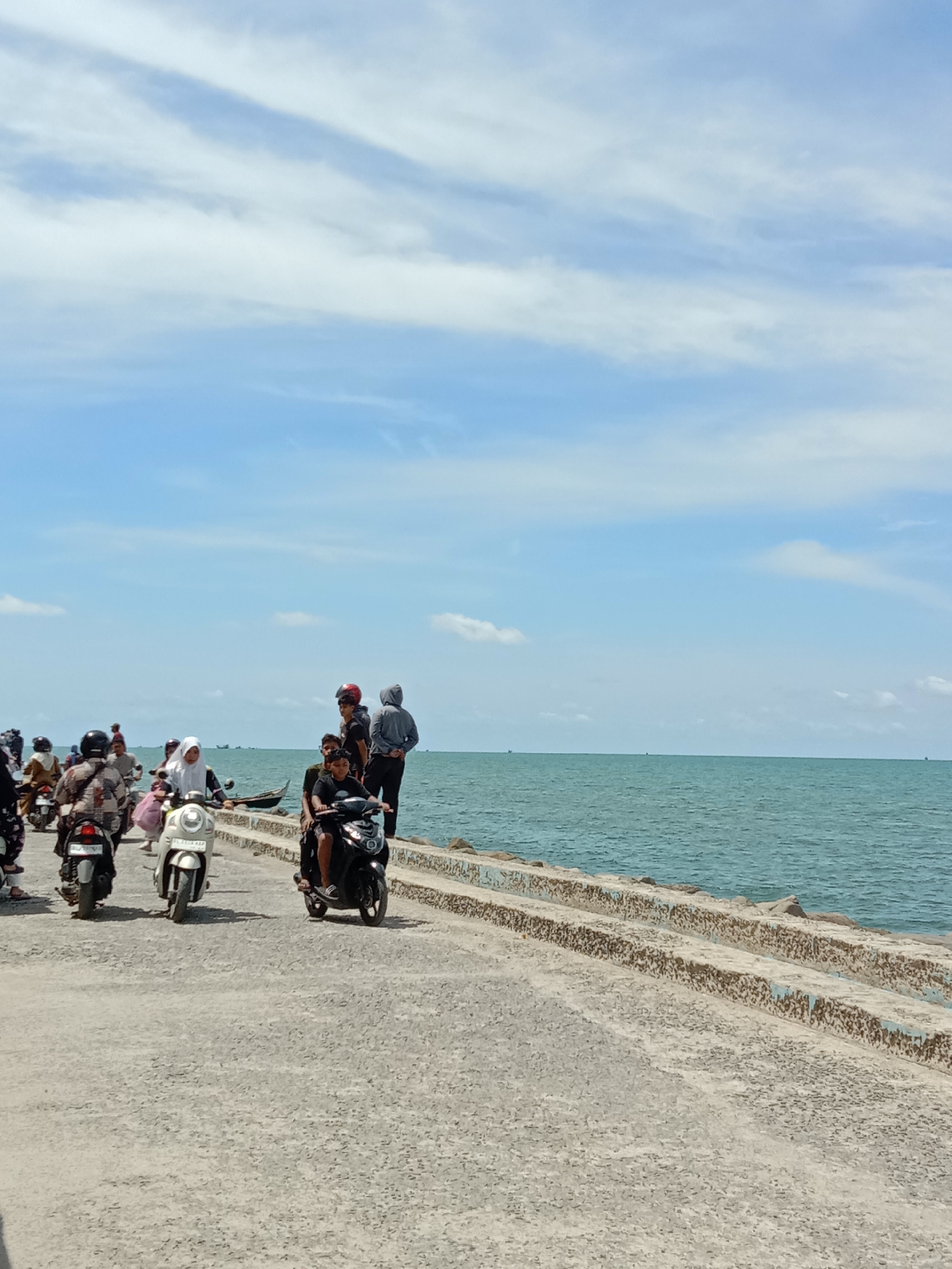 The image shows a coastal scene at Lhokseumawe, Indonesia. A concrete walkway lines the ocean. Several people are gathered, some on motorcycles and scooters. The sky is blue with scattered clouds. The sea is calm with a blue-green tint. In the distance, there are boats on the horizon.