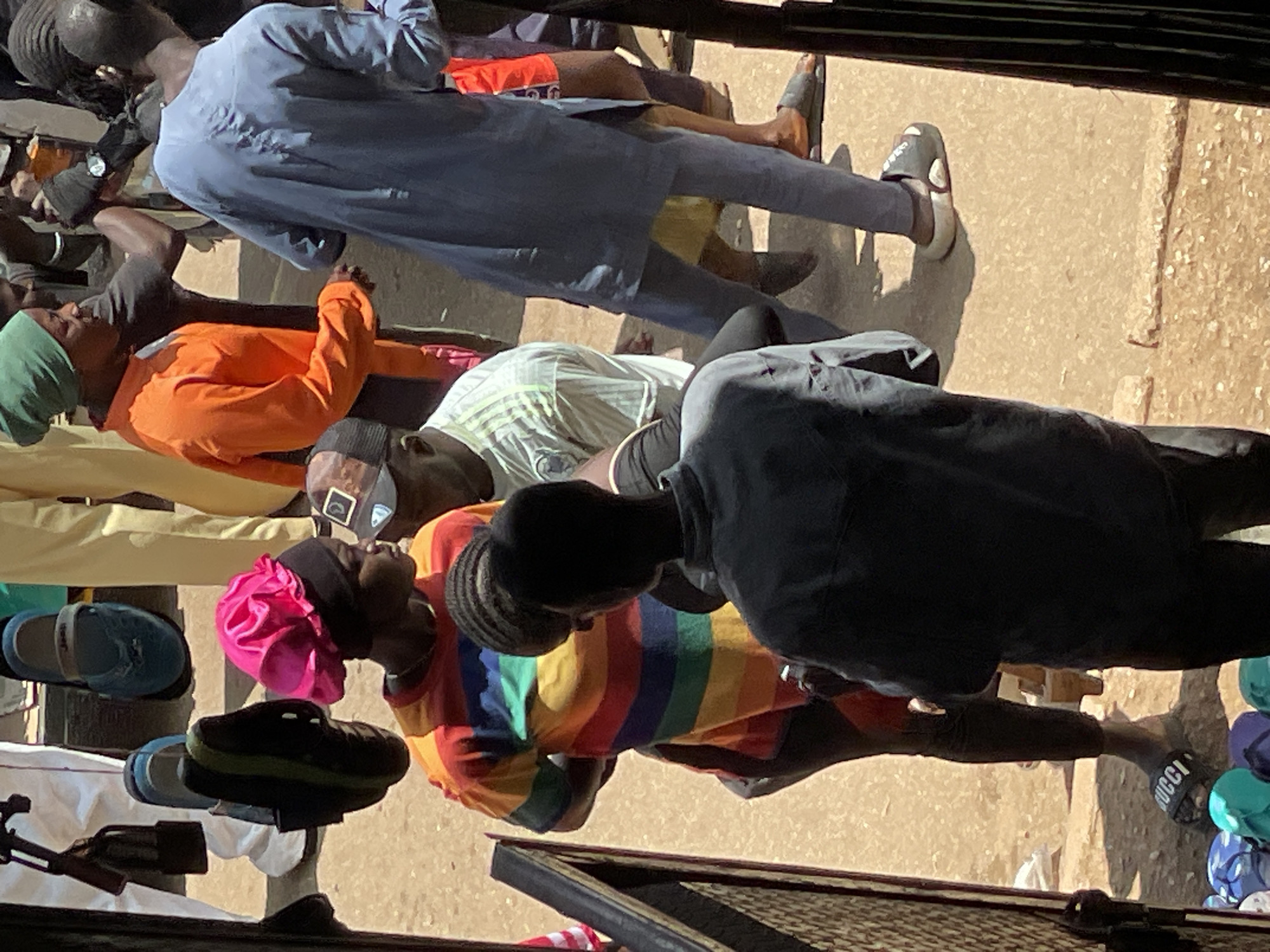 In an outdoor setting in Unknown, Unknown, several individuals are gathered. A person in the foreground is wearing a gray long-sleeved shirt and black pants, with a dark-colored shoe visible on their left foot. To their right, another individual is seen from the waist down, wearing a blue tunic and pants, and a gray sandal on their right foot. Further back, a person in an orange long-sleeved shirt and dark pants is standing. To their left, another person wears a green headscarf and dark clothing. In the middle ground, a person in a rainbow-striped t-shirt and black leggings is visible. A dark-colored shoe is on their left foot. Another individual, wearing a white t-shirt with a green trim and a baseball cap, is positioned behind them. Various shoes, including sandals and flip-flops, are scattered on the ground in the lower portion of the image. The ground appears to be made of sand or dirt.