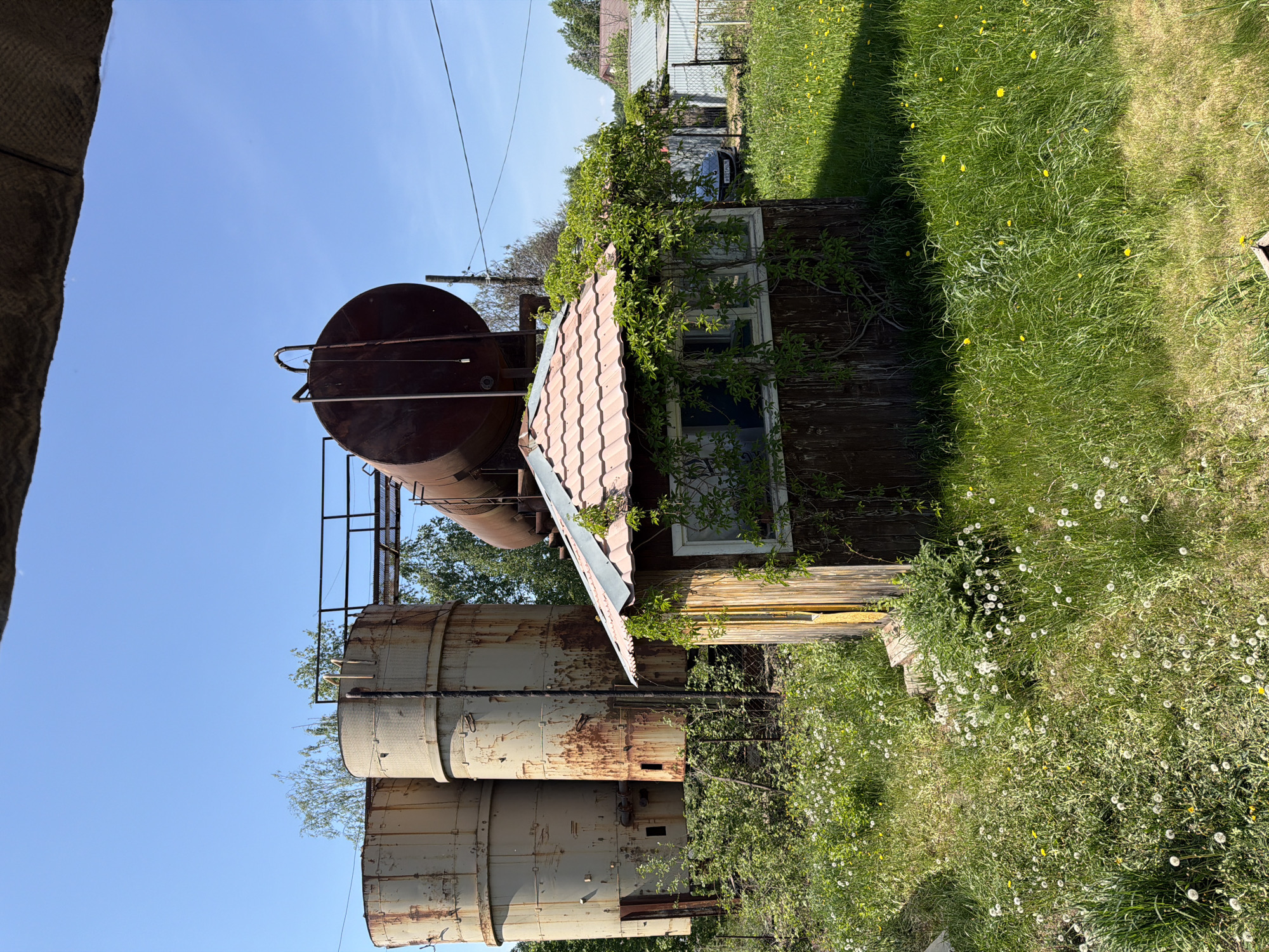 The image shows a dilapidated structure located in Ягул, Russia. Three large, cylindrical, rusty metal tanks stand vertically, topped with a larger horizontal tank. A small, weathered wooden building is situated nearby, partially overgrown with vegetation. A rusted, red-tiled roof covers part of the building. The surrounding environment consists of overgrown grass and weeds, with a glimpse of other buildings and a chain link fence in the background. The sky is clear and blue. No people are visible.