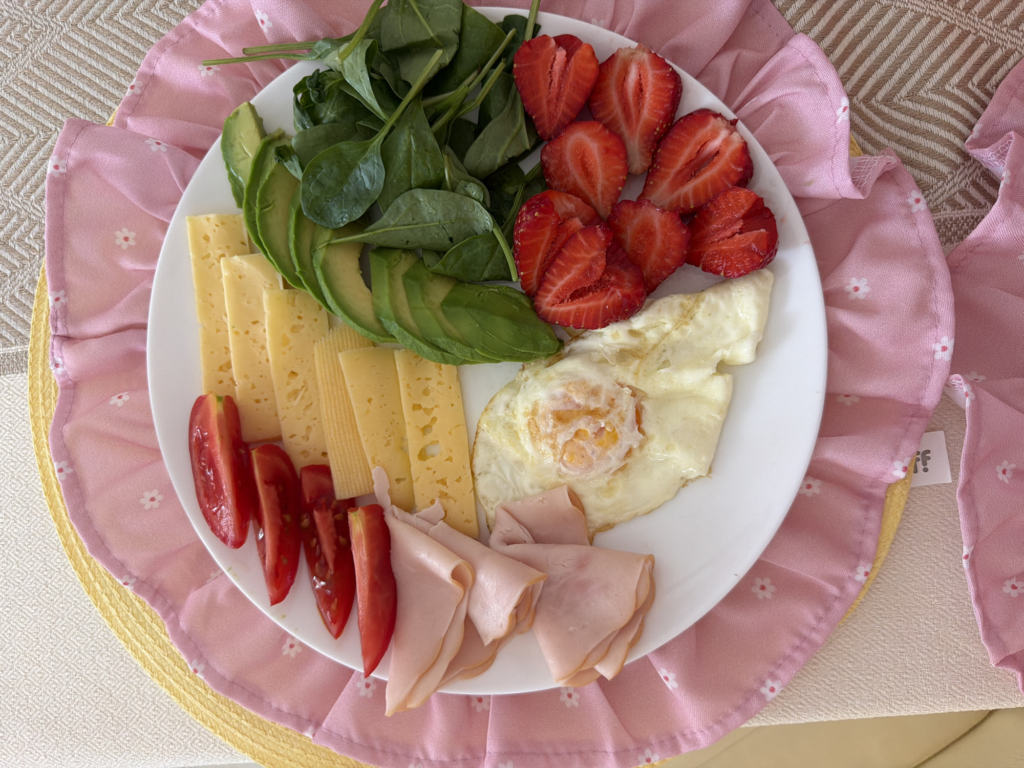 A white plate holding a meal is placed on a pink and yellow placemat. The meal consists of sliced tomatoes, sliced cheese, sliced avocado, spinach, sliced strawberries, and a fried egg. The plate is centrally located on the placemat. The placemat has a ruffled edge with a floral pattern. The placemat is on top of a textured surface that appears to be a table or tablecloth. The background is a neutral-colored couch or chair. There are no people or animals visible in the image. The setting appears to be a domestic indoor environment. There is no information in the image to indicate a location in Moscow, Russia.