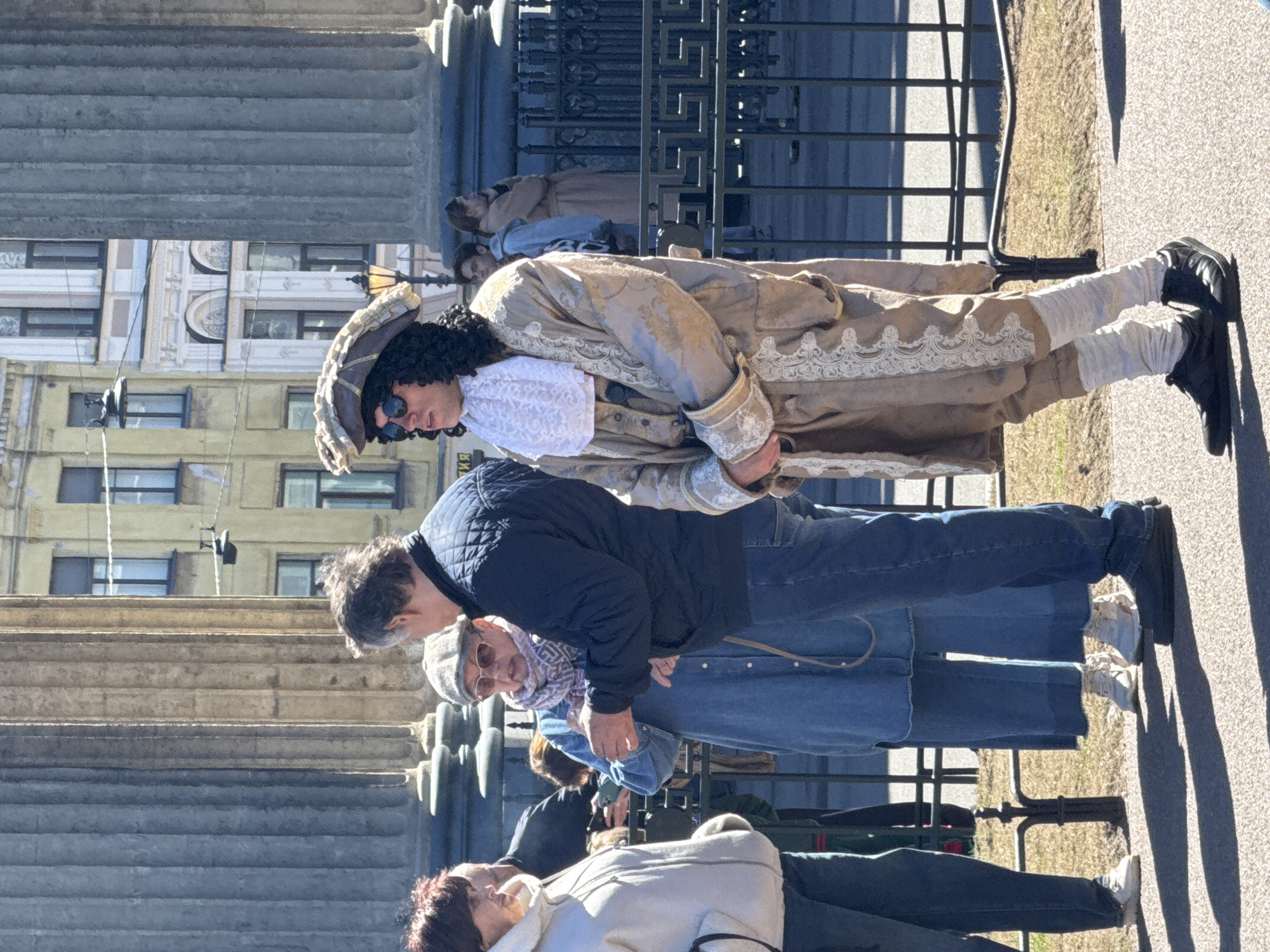 The image shows people in the city of Saint Petersburg, Russia. A person dressed in an 18th-century costume, wearing sunglasses, and a wig, stands in front of a group of people. The background features architectural structures with columns and windows, typical of Saint Petersburg. Fencing is visible between the person in costume and the group. The setting is outdoors on a paved surface, under daylight.