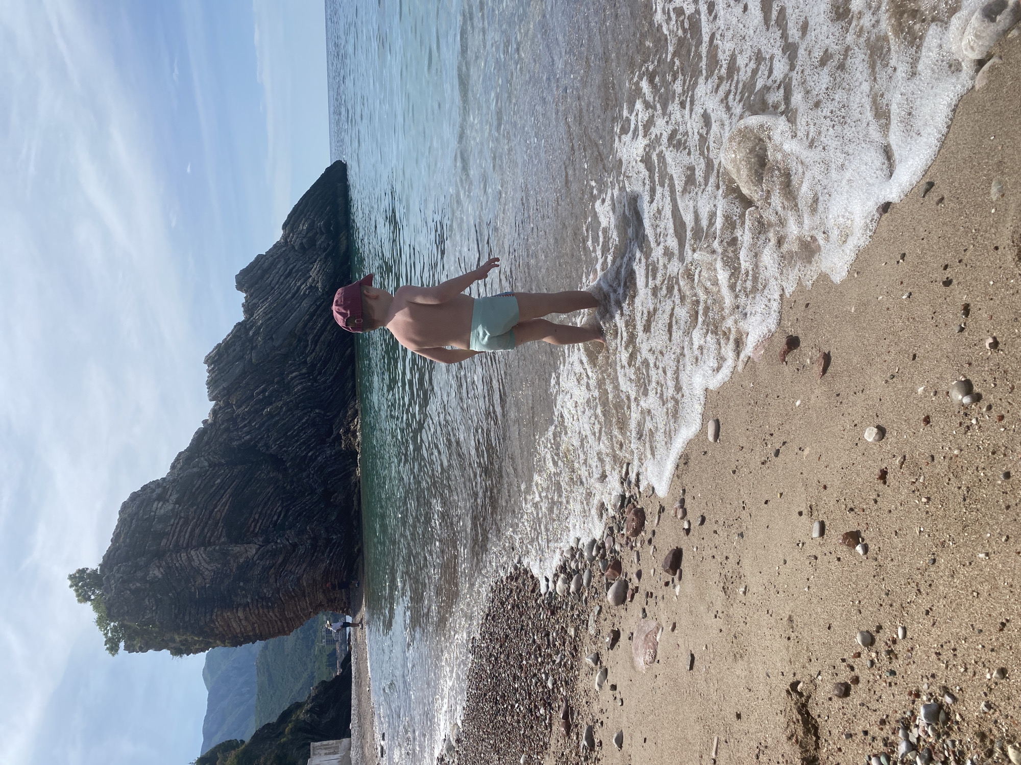 A sunlit, rotated image shows a child in the water on a beach in Rafailovići, Montenegro. The child wears a maroon baseball cap and light green swim trunks. The child stands facing the water with arms outstretched. To the left is a large, dark rocky outcrop, and behind it, distant mountains. The water's edge is foamy. The beach is composed of both fine sand and small pebbles. Other individuals can be seen in the background near the rocks. The sky is light blue with wispy clouds.