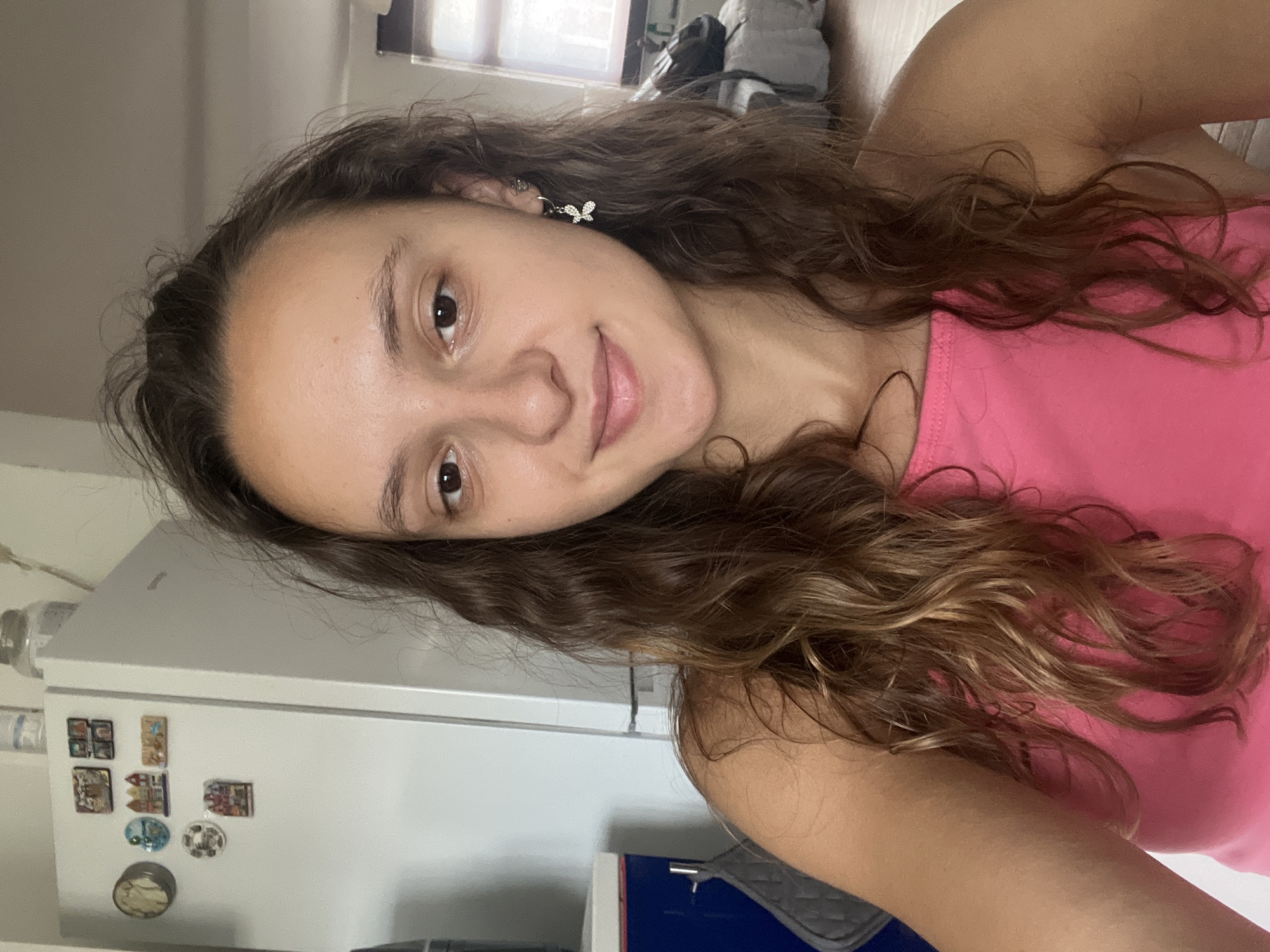A young woman with medium-length, wavy brown hair and brown eyes is pictured in a close-up selfie. She is wearing a bright pink tank top and a small silver earring in the shape of a butterfly. Her skin appears clear and her expression is neutral.
The background suggests an indoor setting, possibly a kitchen or living area. To the left, a white refrigerator is visible, adorned with various small magnets, including what appear to be miniature buildings. A dark blue cabinet with a silver handle is also present to the left. The right side of the image shows a window with diffused natural light and a bed or sofa with grey and white bedding, and a dark bag on it. The general environment appears to be a residential space.
This image was taken in the city of Saarbrücken, Germany.