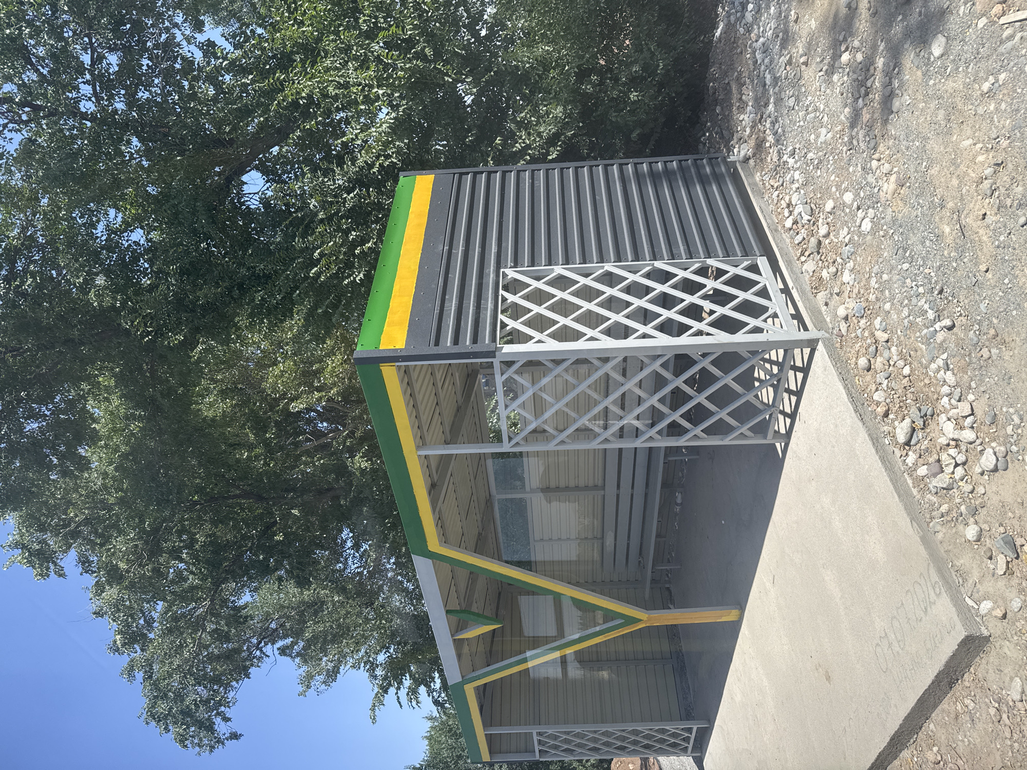 An outdoor bus stop structure is visible in Кашка-Суу, Kyrgyzstan. The structure has a metal roof with green and yellow stripes. The walls are made of corrugated metal in a dark gray color. There is a white lattice-like screen on the left side of the bus stop, and a bench inside. A concrete pad extends from the bus stop, with gravel and some rocks surrounding it. Trees with green foliage are in the background, and the sky is clear and blue. The concrete pad has some faint markings that appear to be writing.