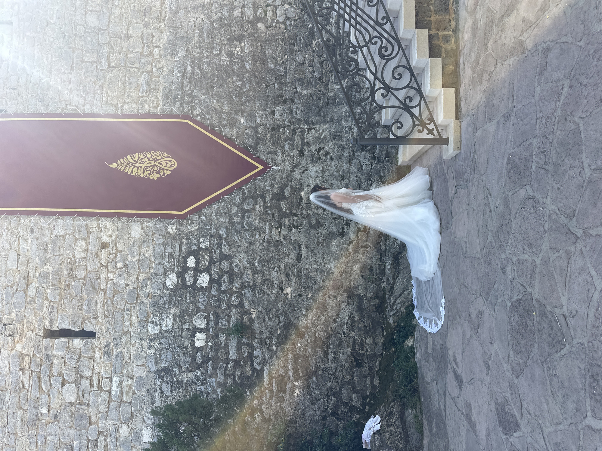 The image shows a woman in a white wedding dress and veil standing on a stone walkway next to a stone wall with a burgundy banner. The location is near stairs with a black iron railing, possibly in or near Budva, Montenegro. The lighting suggests daylight conditions. The composition includes architectural elements typical of the region.