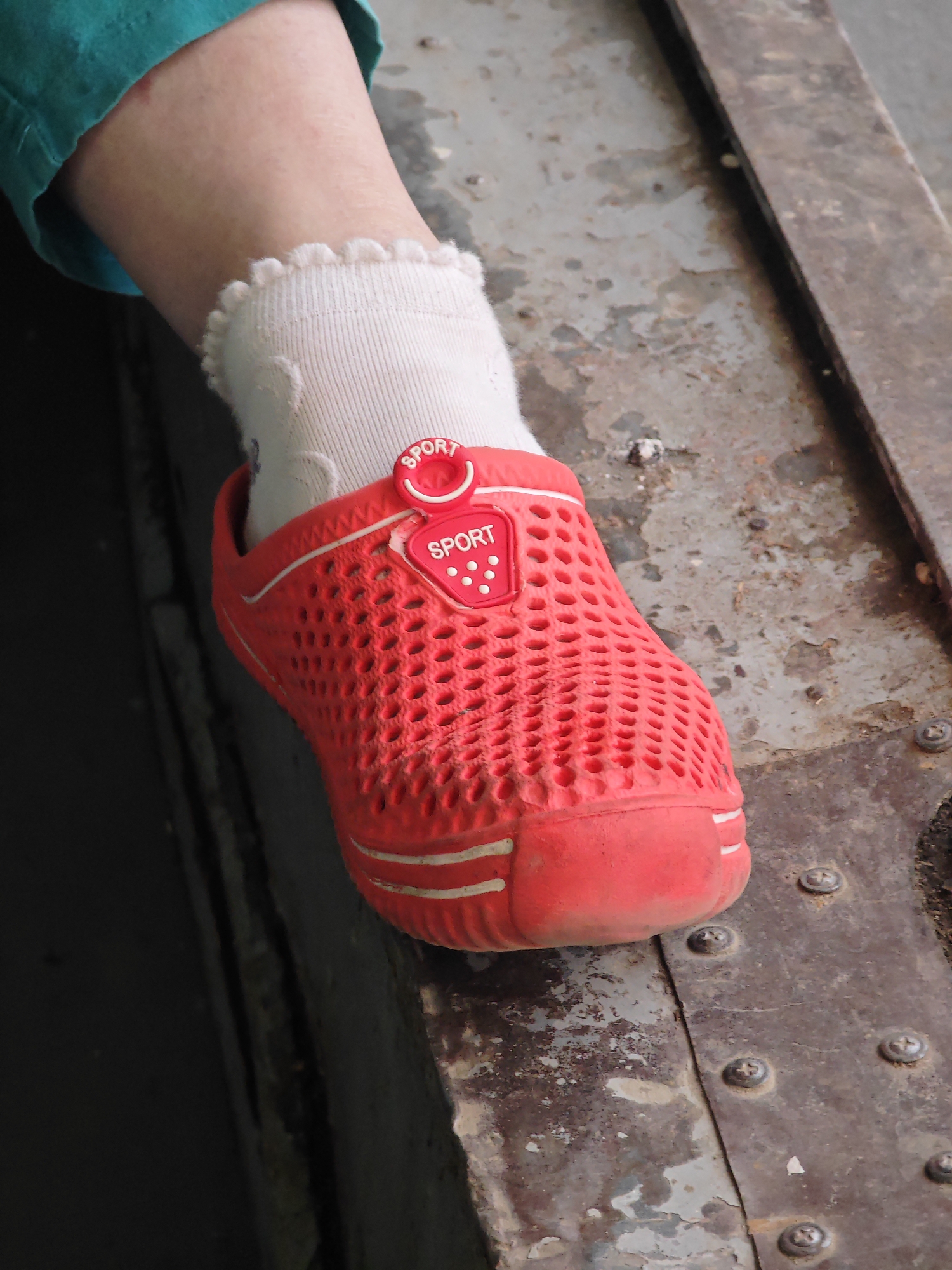 A close-up, eye-level shot focuses on the right foot of an unseen person, wearing a red clog-style shoe with white trim and white socks with a ruffled edge. The shoe has a decorative red tag with the word "SPORT" in white lettering. The foot is resting on a weathered, rusty metal surface. The background includes a dark, out-of-focus area and a portion of the person's leg covered by a teal garment. The image does not provide any information that can be used to determine its location within the city of Krasnoyarsk, Russia.