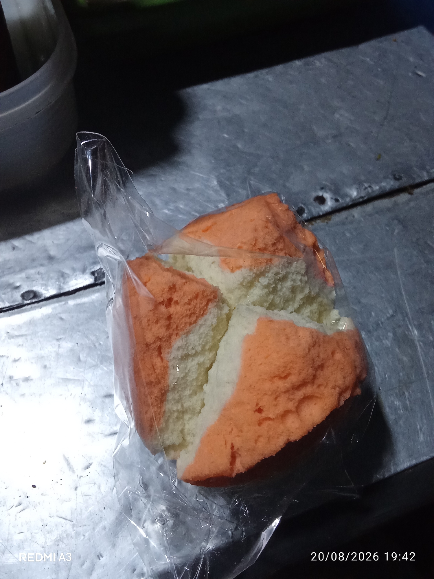 The image displays a sliced, two-toned cake, primarily orange and white, encased in clear plastic packaging. The cake exhibits a fluffy, airy texture with visible air pockets. It rests on a metallic surface, possibly a table or counter, which appears scratched and worn. In the background, a dark, out-of-focus object is visible on the left, and to the right, the metallic surface continues. A timestamp in the lower right corner indicates "20/08/2026 19:42," and "REDMI A3" is faintly visible in the lower left corner, suggesting the device used for capture. The lighting is dim, creating shadows and highlighting the texture of the cake and the surface it's on. There are no people or animals present in the scene. The overall impression is a casual, perhaps food-stall or home-based setting, with the focus solely on the packaged cake. The location in Cemani, Indonesia, is inferred from the context provided, but not visually discernible from the image itself.