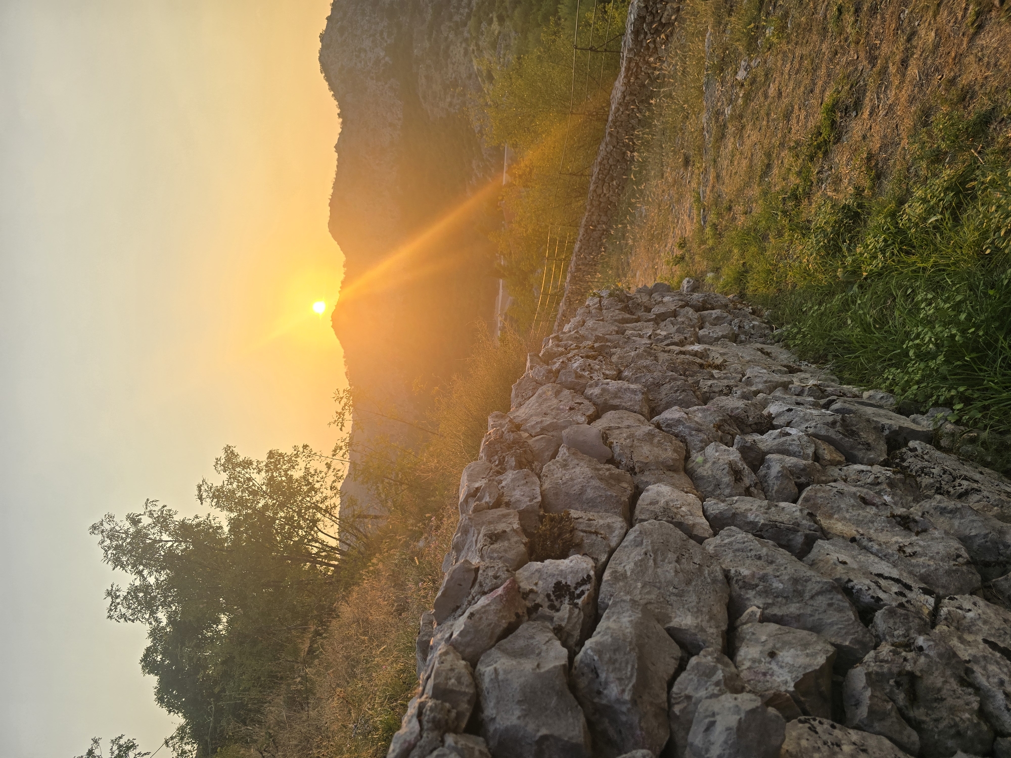 The image depicts a sunset scene with a low-lying sun casting a warm orange glow over a mountainous landscape. In the foreground, a textured stone wall runs diagonally from the lower left to the mid-right of the frame, constructed from irregular grey stones. Adjacent to the wall, dry, golden grass and some green vegetation cover the ground. Further back, a path is visible, bordered by a low stone wall and a metal railing, leading towards a valley. The valley contains scattered trees and shrubs, also bathed in the golden light. The background features rolling hills and mountains silhouetted against the bright sky, with the sun positioned just above the horizon, emitting strong sunbeams. The sky transitions from a pale yellow near the sun to a lighter hue towards the top. The overall impression is one of a natural, rustic environment during golden hour, likely in or around Gornja Vrbica, Montenegro, given the mountainous terrain and stonework common in the region. No people or animals are visible in the scene.